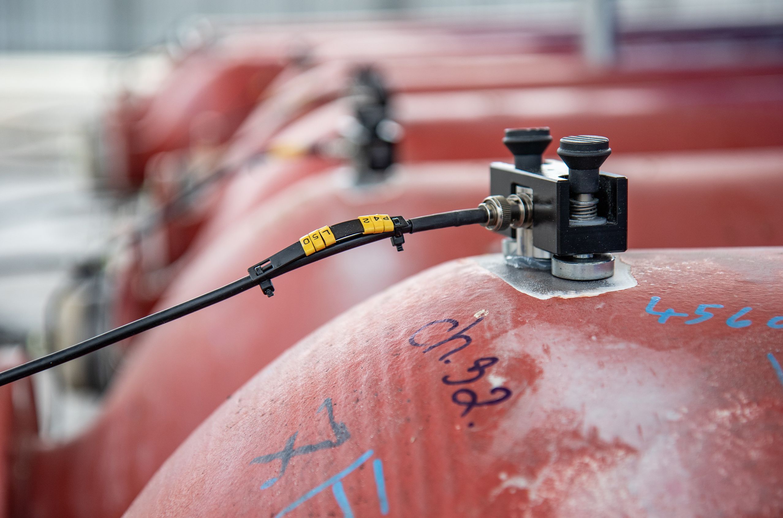 Close-up of a cable and connector on a red industrial tank, with blue markings and several similar tanks in the blurred background.