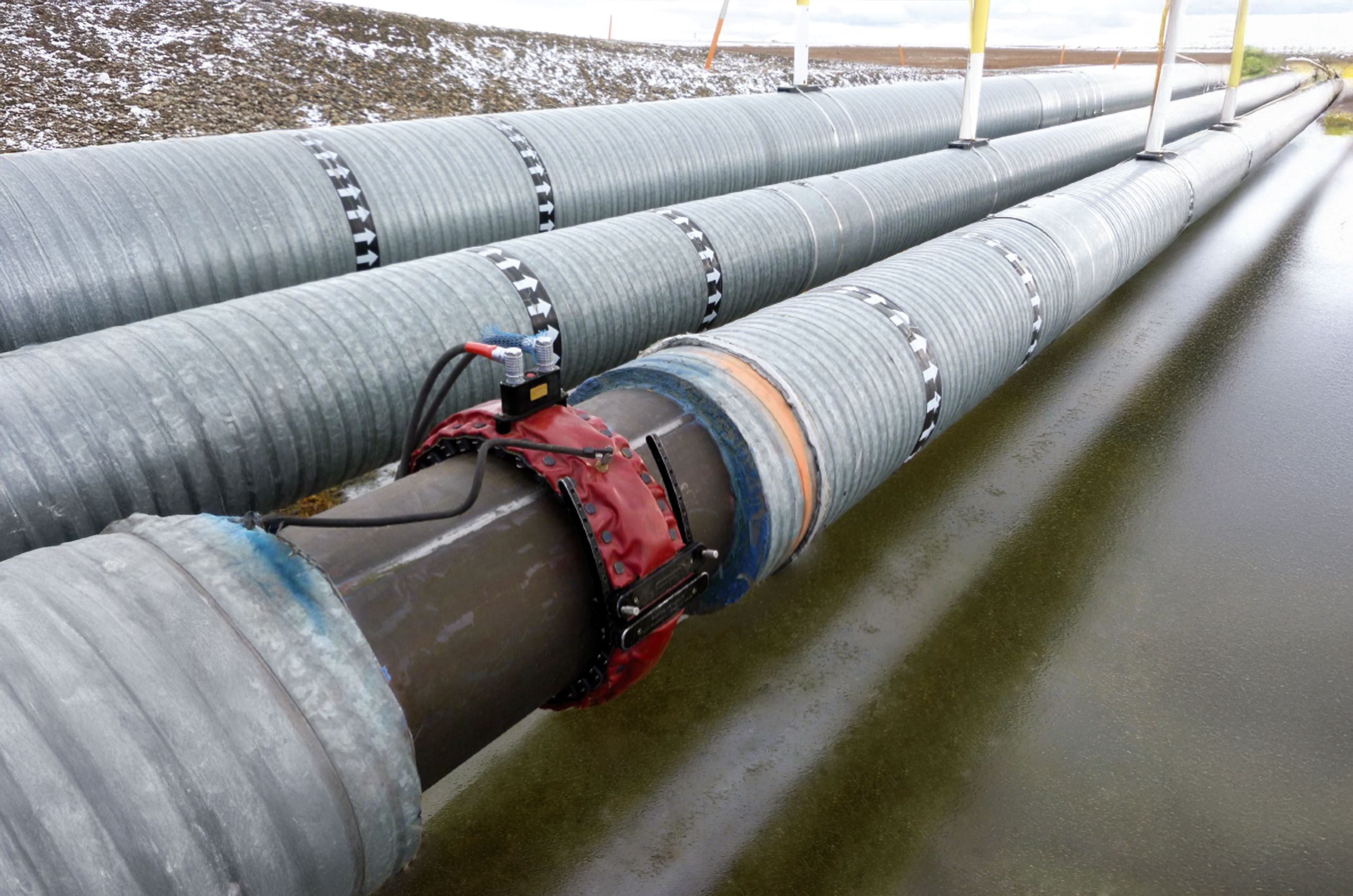 Metal pipelines with sensors run parallel, elevated above water, surrounded by a snowy landscape.