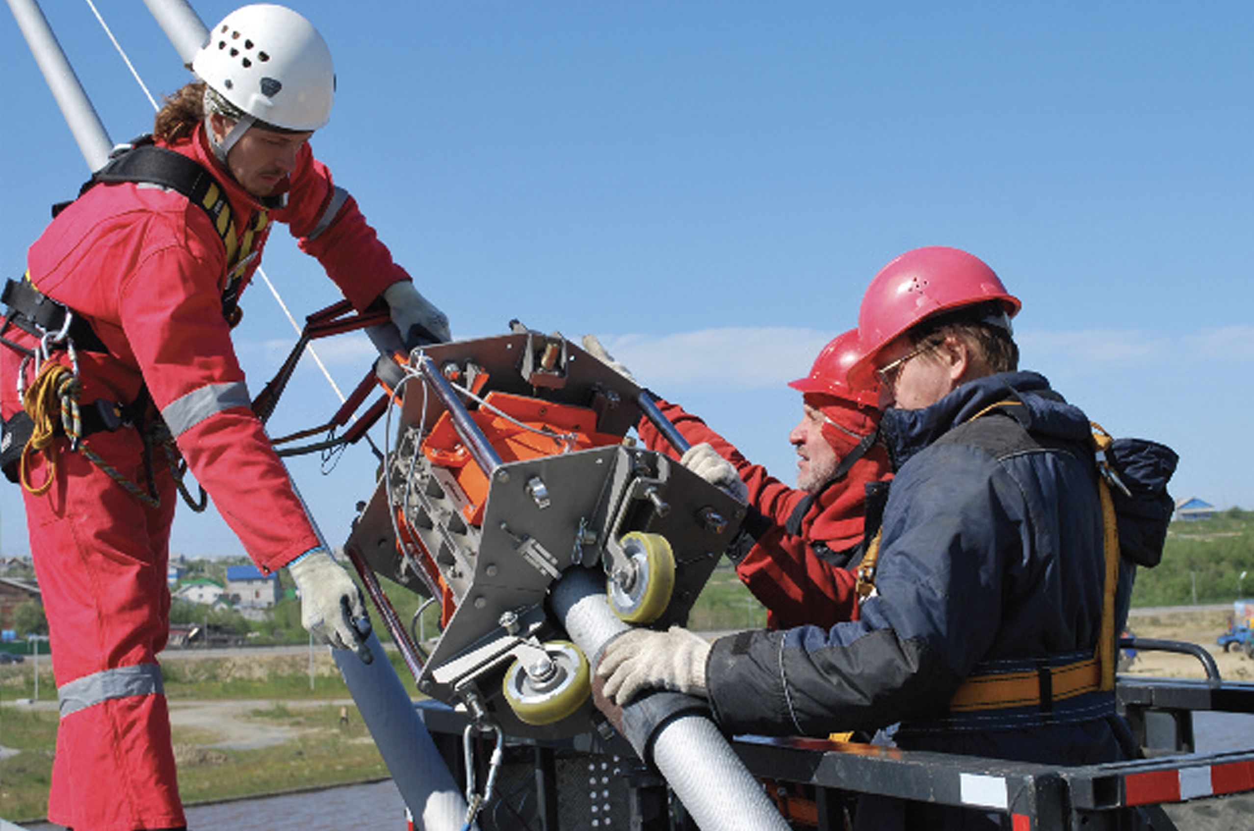 Three workers in safety gear operate machinery on a high platform, with a clear blue sky and landscape in the background.