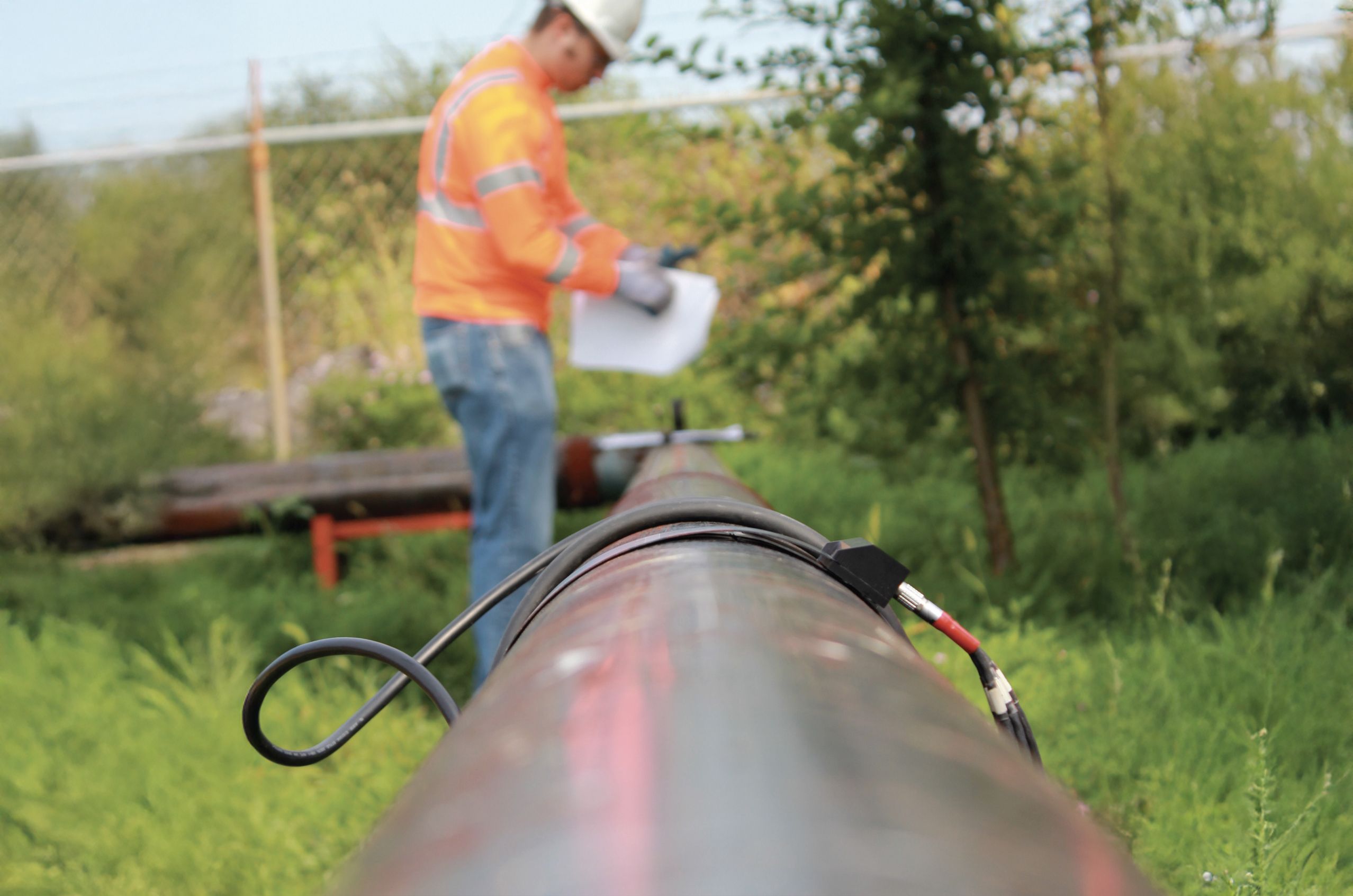 Worker in an orange vest and helmet inspects a pipeline with cables attached, surrounded by greenery.