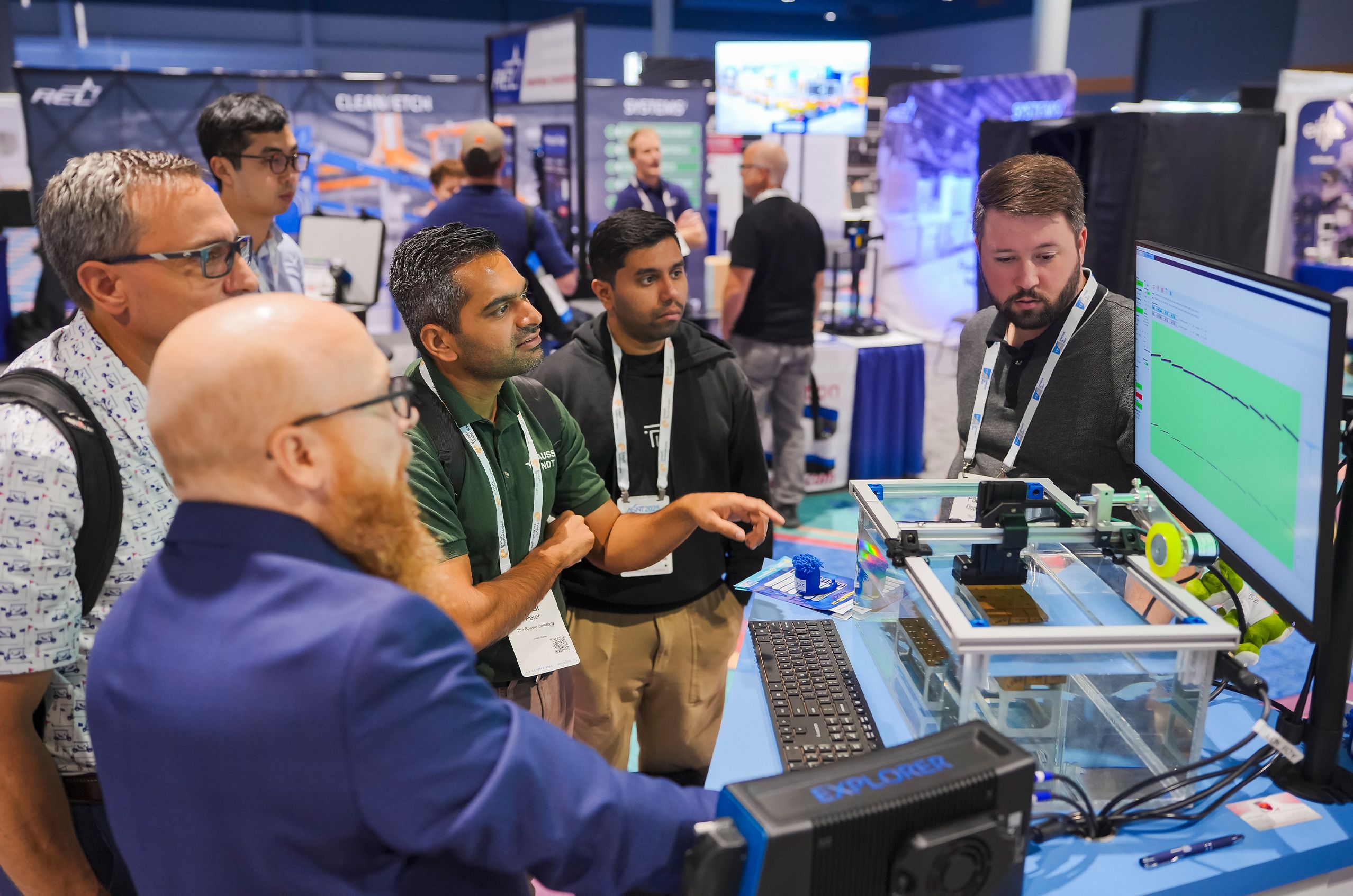 A group of men attentively observe a technology demonstration at a trade show booth, featuring a screen and hardware setup on a table.