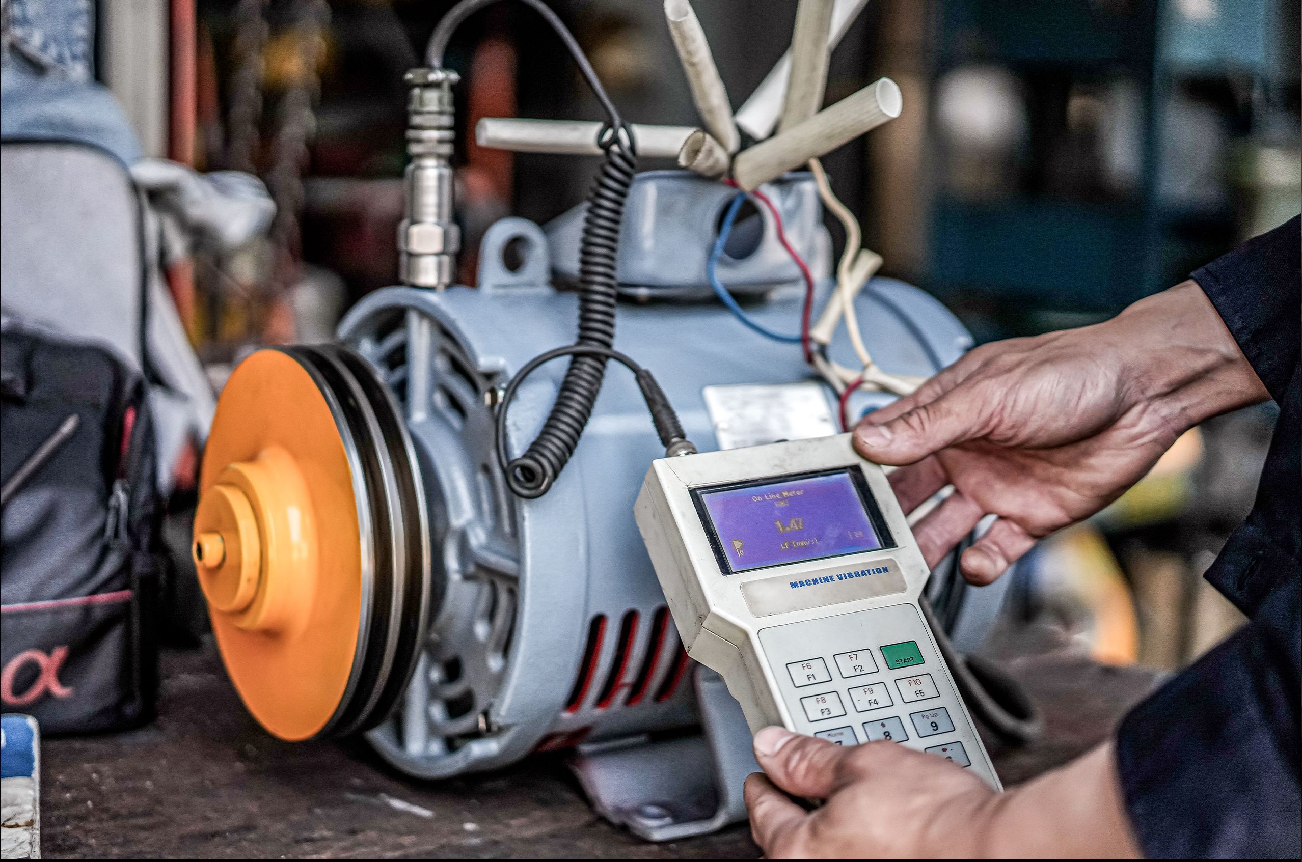 Hands using a machine vibration meter to test a motor, with wires and a yellow pulley visible.