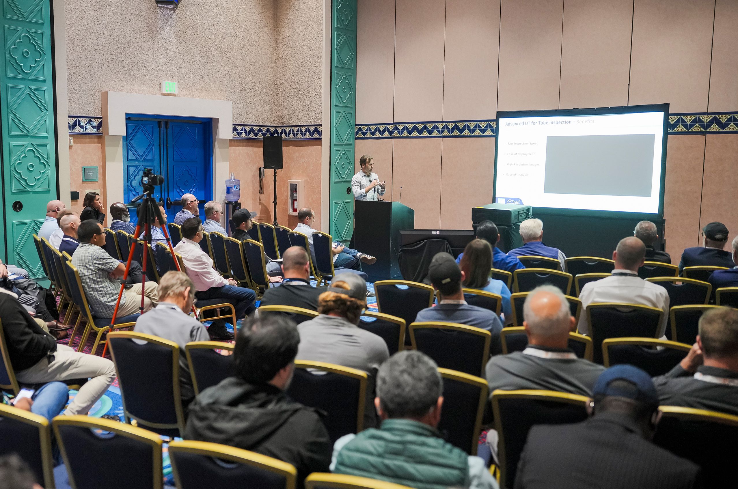 A speaker presents to a seated audience in a conference room, with a large screen displaying a presentation.