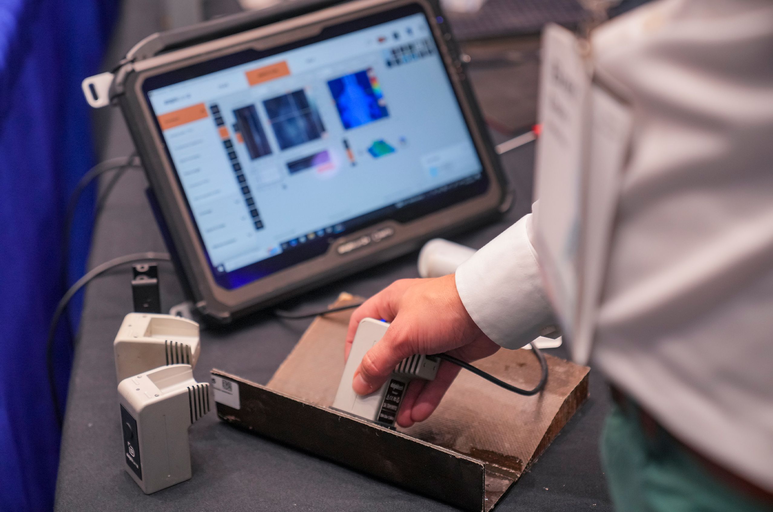 A person holding a handheld ultrasonic testing device over a metal surface, with a tablet displaying images and data in the background.