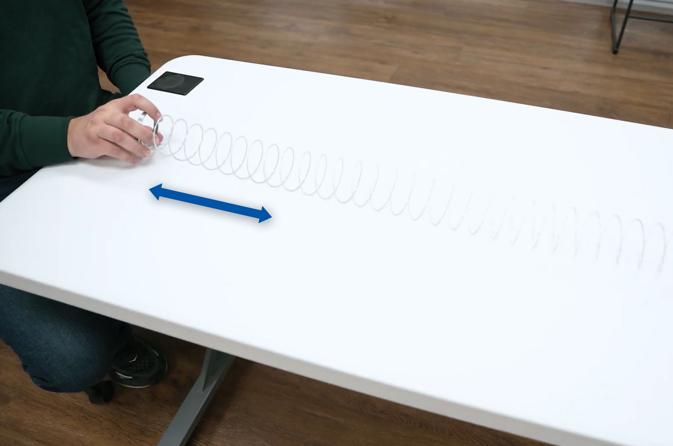 Person demonstrating wave motion with a slinky on a white table, showing a blue arrow indicating direction.