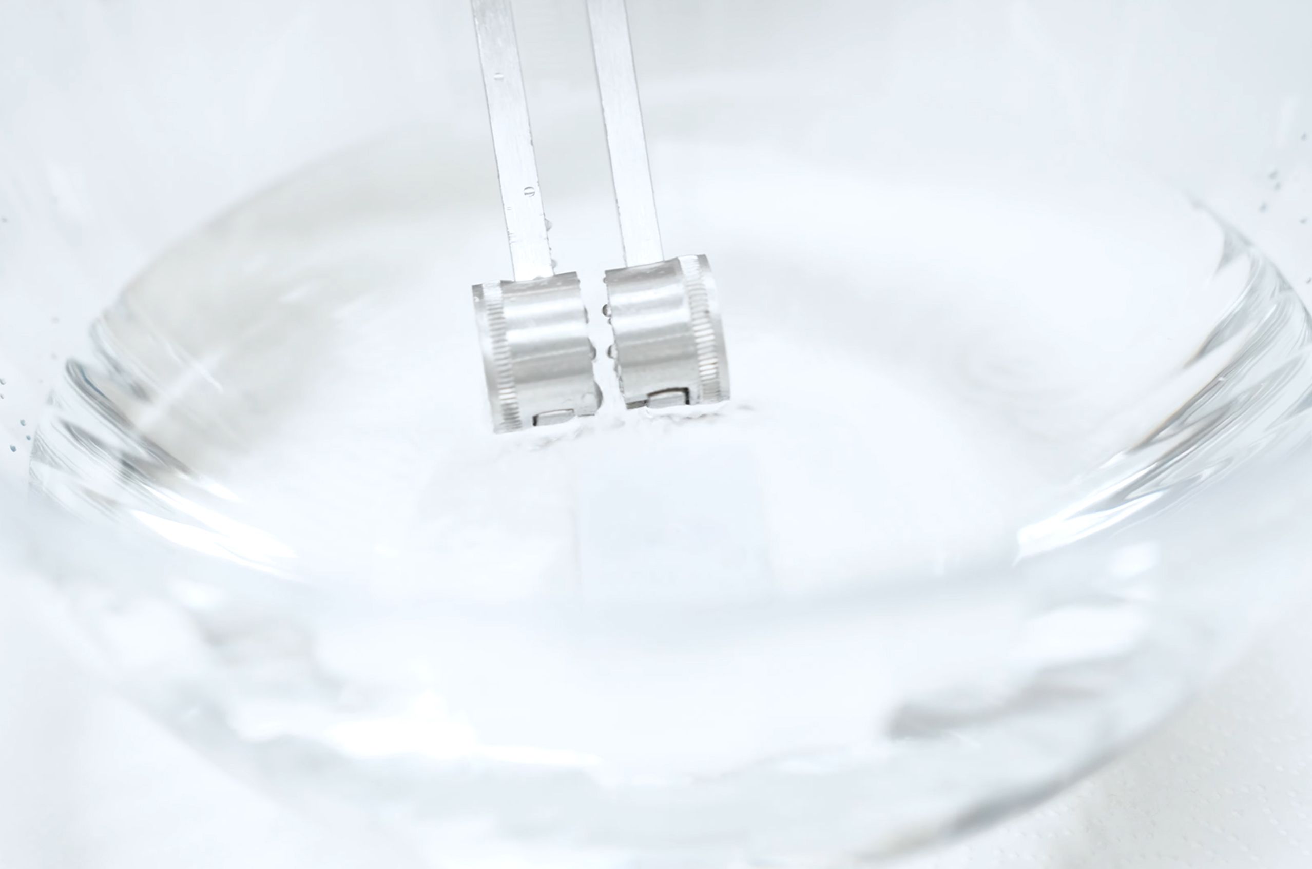 Close-up of water with two metal prongs of a tuning fork partially submerged, creating waves in the water.