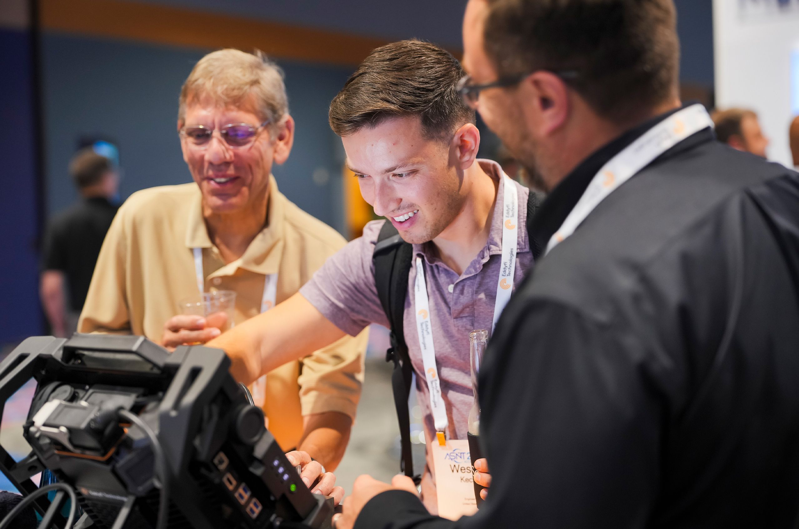 Three men interact with a tech device at a ASNT 2025. One points at the screen, while the others watch, smiling.