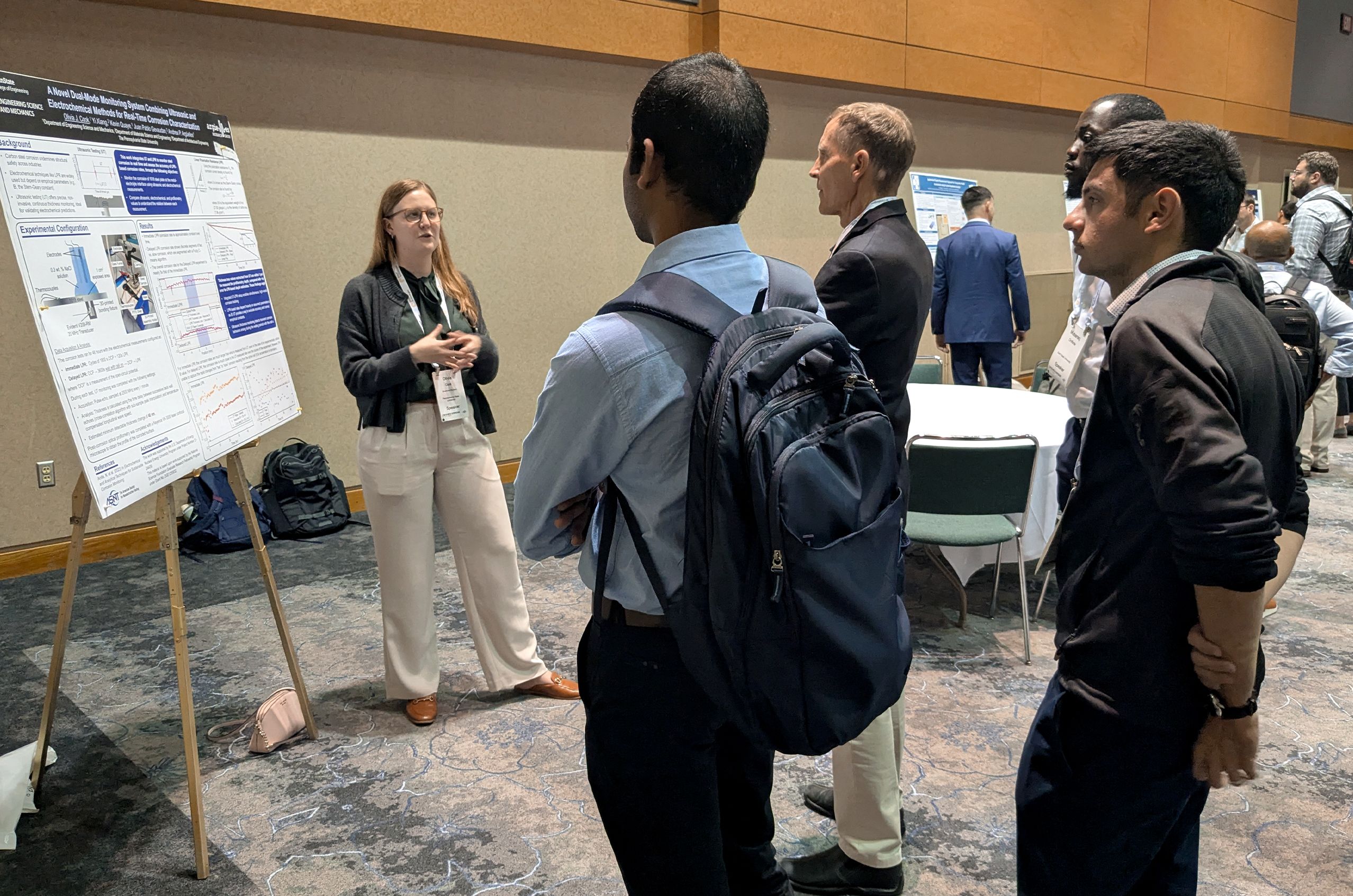 A woman presents a research poster to four attentive men in a conference setting. They are gathered around her, discussing the content.