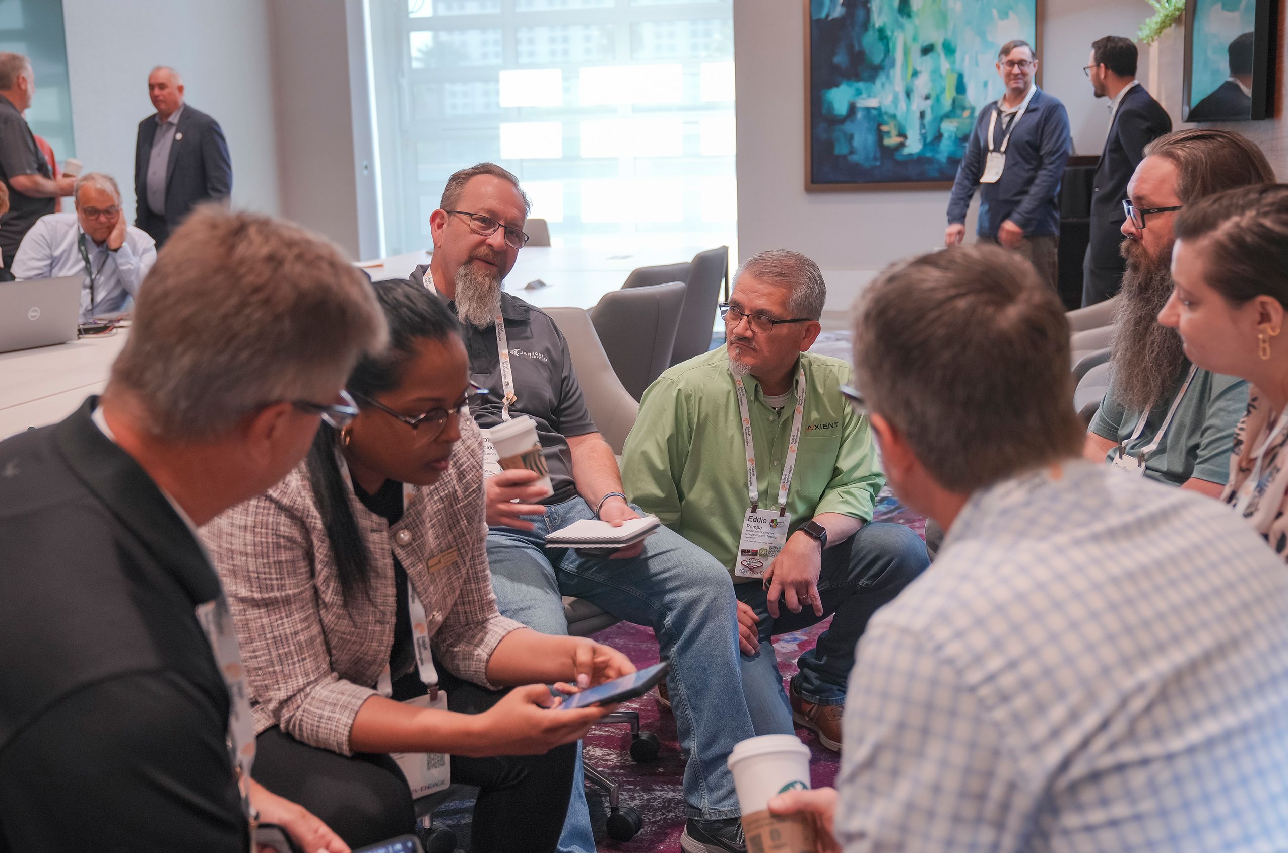 A group of people seated in a circle, engaged in discussion, some holding coffee cups. A modern office setting with a large window and artwork.