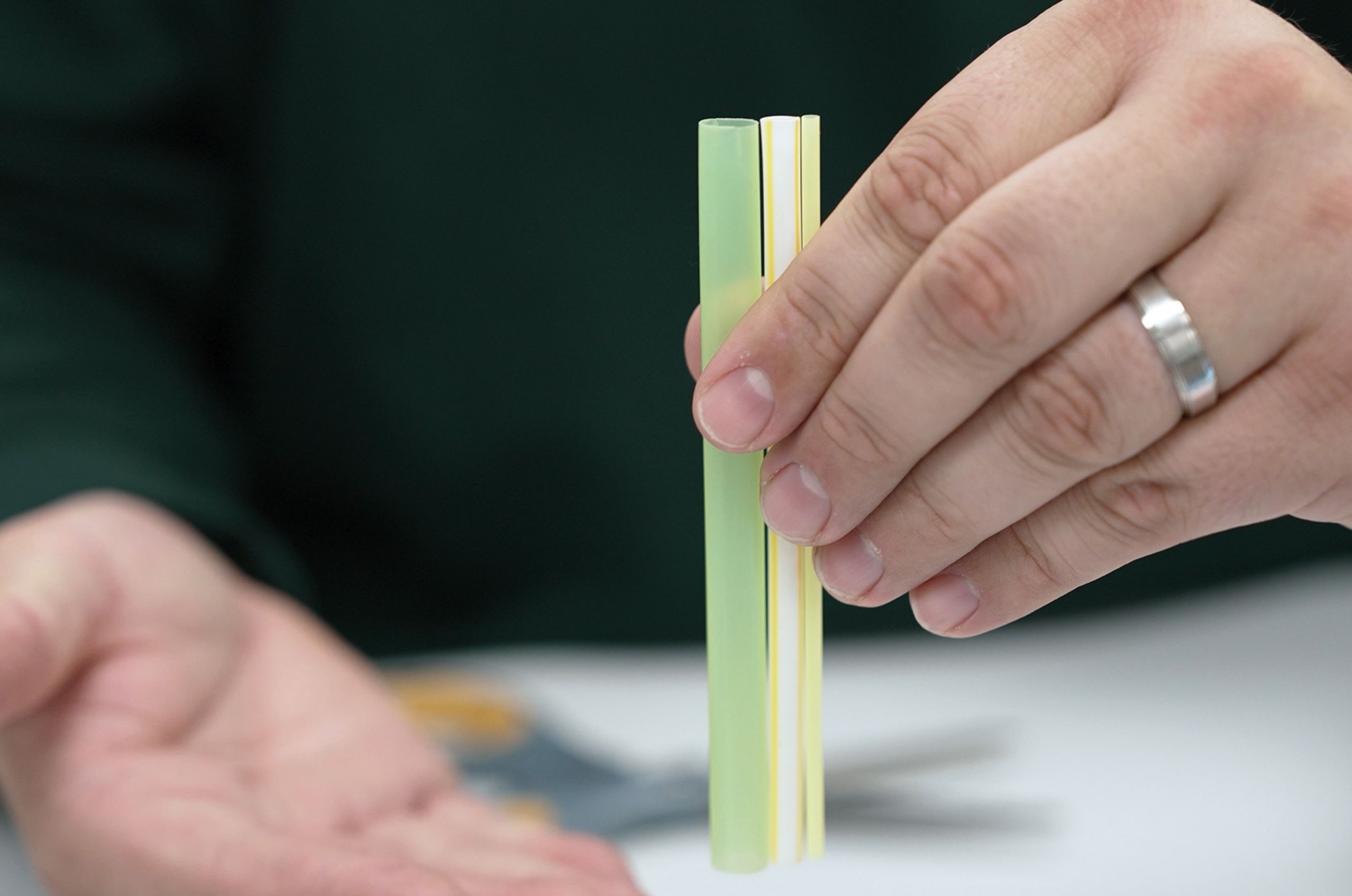 A hand holding three pastel-colored straws, two green and one yellow, with a pair of scissors in the background.