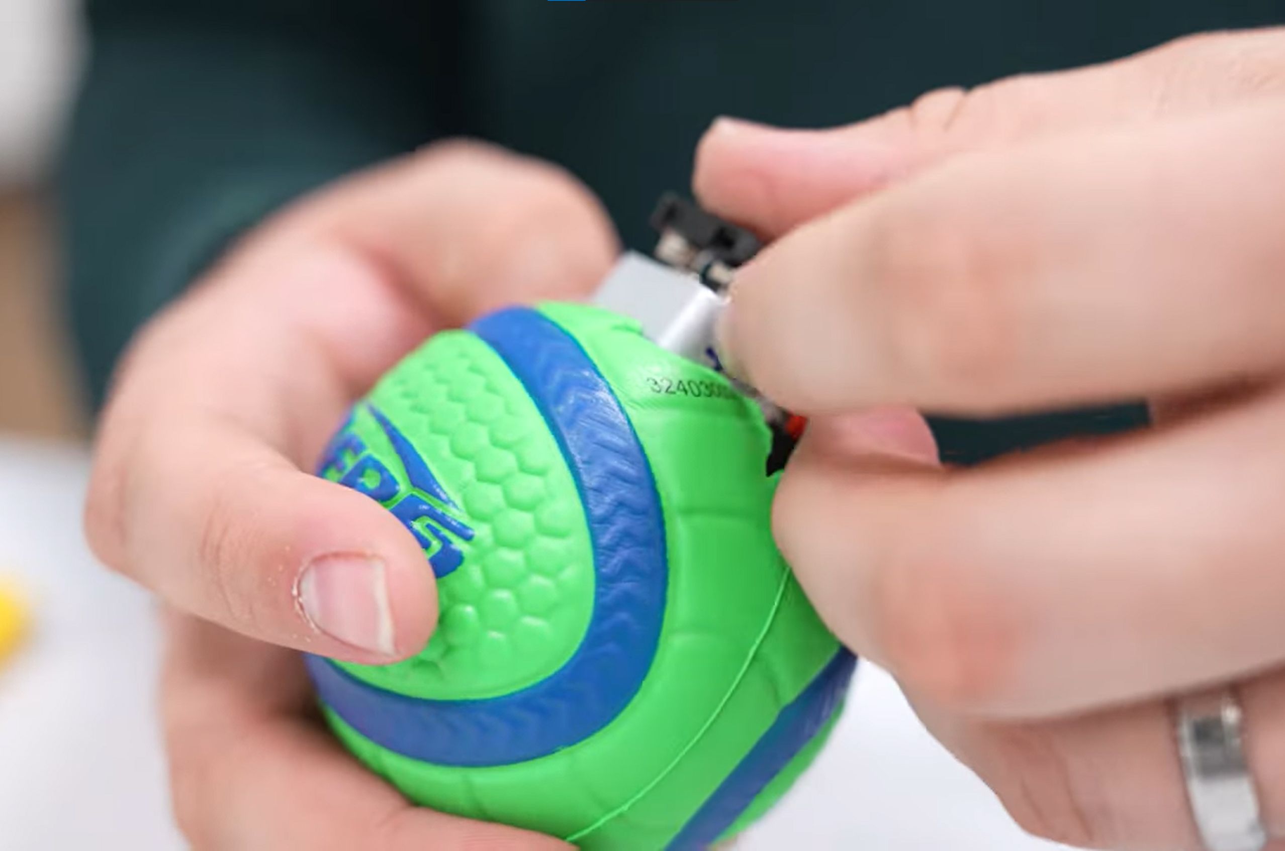 Hands holding a green and blue textured toy ball with a small hole. The hands are pressing in a battery and wires into the ball.