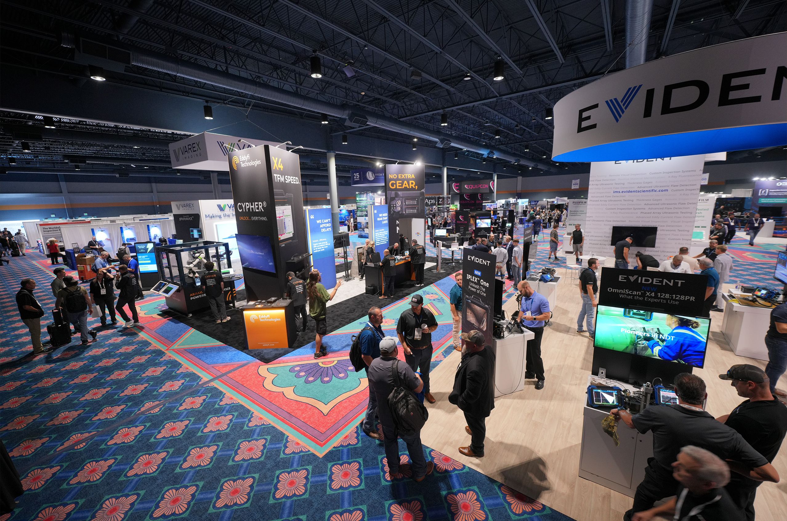 Wide-angle view of the ASNT 2025 exhibition hall with attendees exploring various booths and displays under bright overhead lighting.