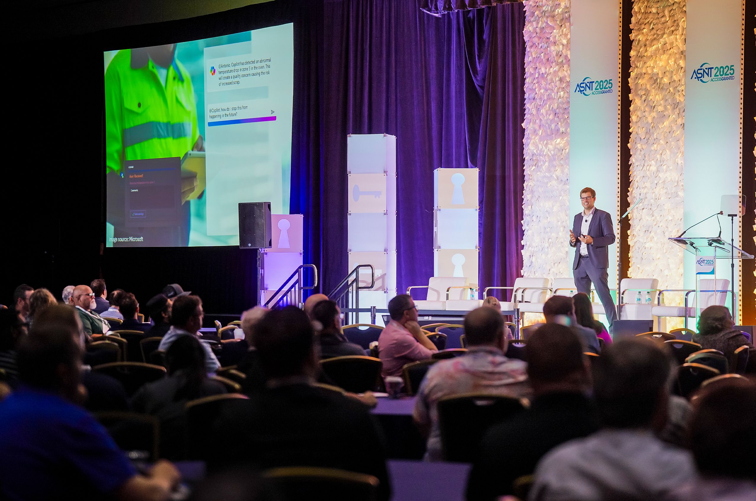 A speaker presents on stage at a conference, with an audience seated. A large screen displays a colorful presentation slide.