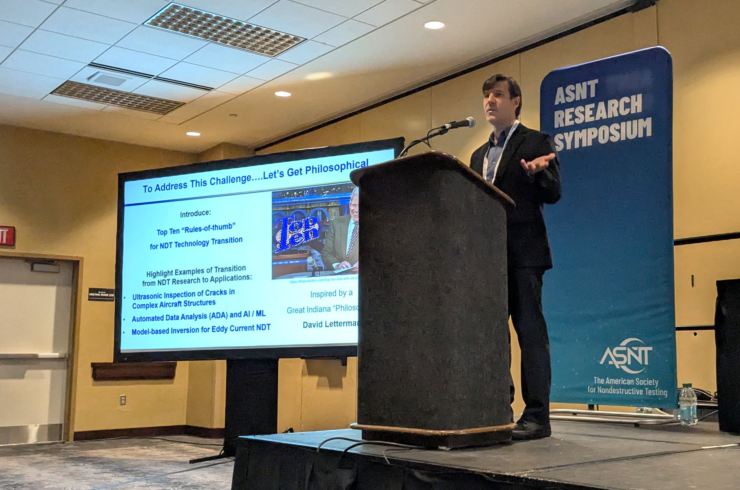 A man stands at a podium presenting at the ASNT Research Symposium. A screen displays a slide with text.