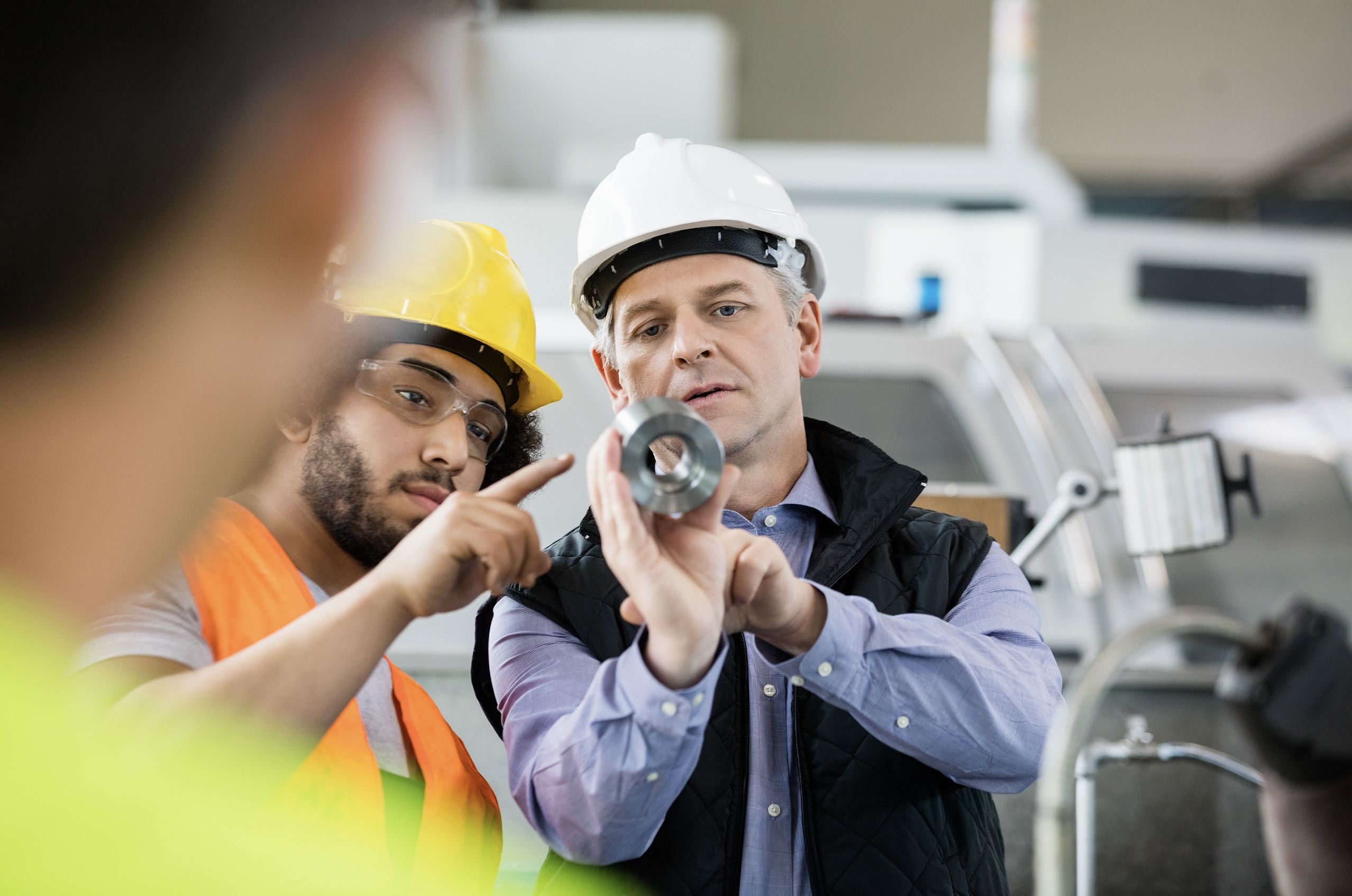 Two workers in safety gear examine a metal part in a factory setting, discussing its features. Machinery is visible in the background.