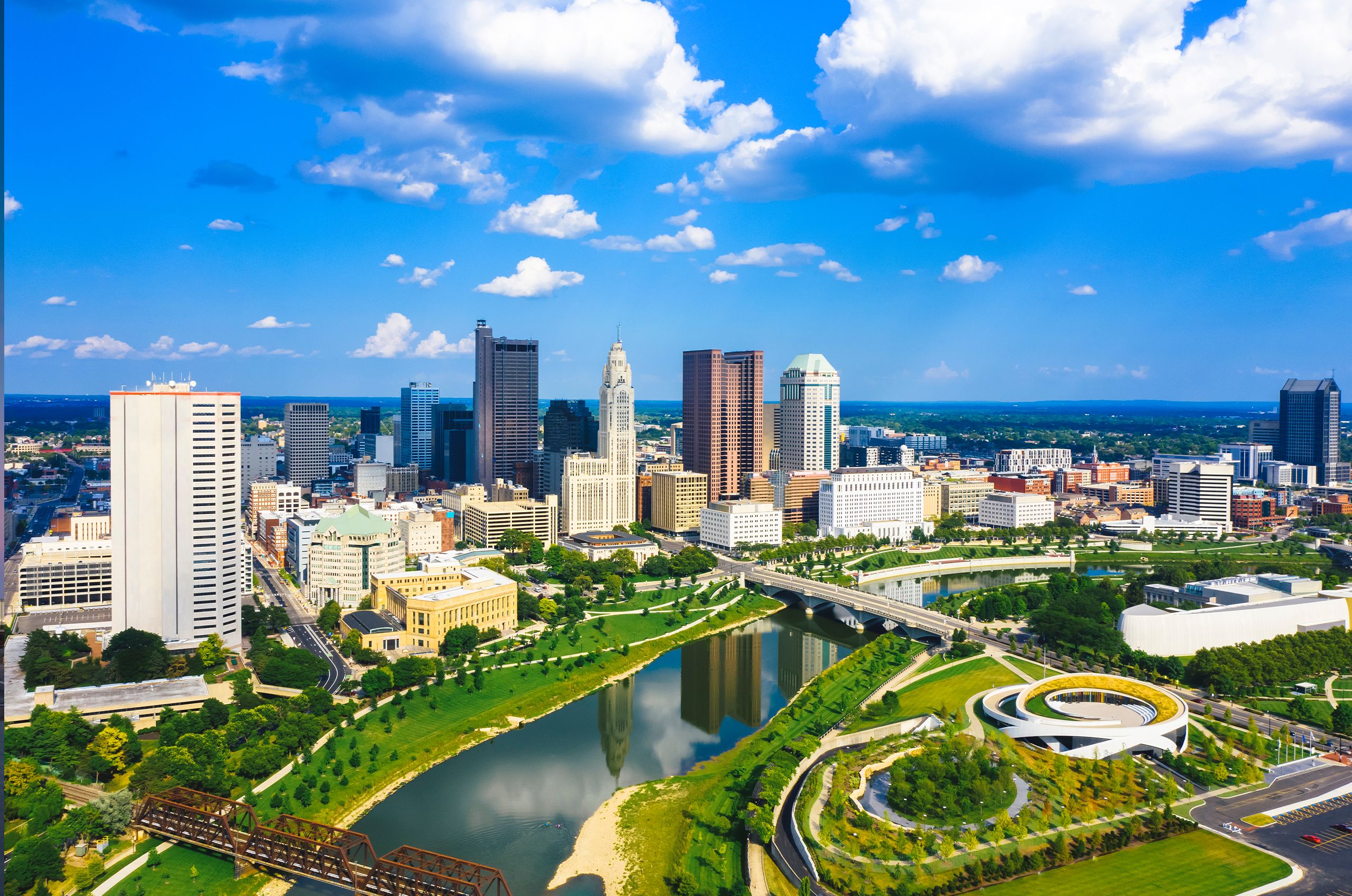 Aerial view of Columbus with tall buildings, a river, and green spaces under a blue sky with clouds.
