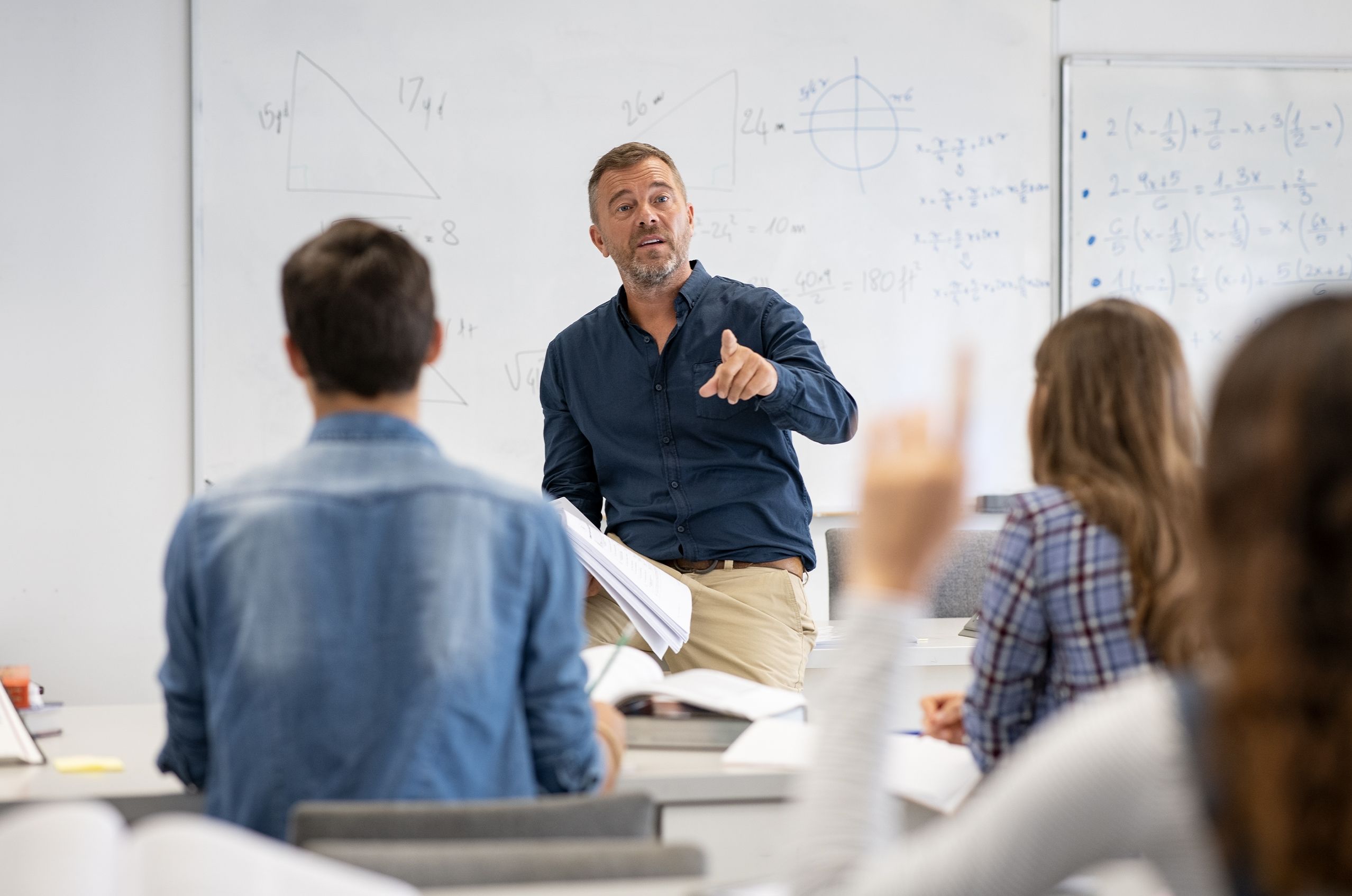 A teacher in a classroom gestures while speaking to students. Geometric diagrams and equations are drawn on the whiteboard behind him.