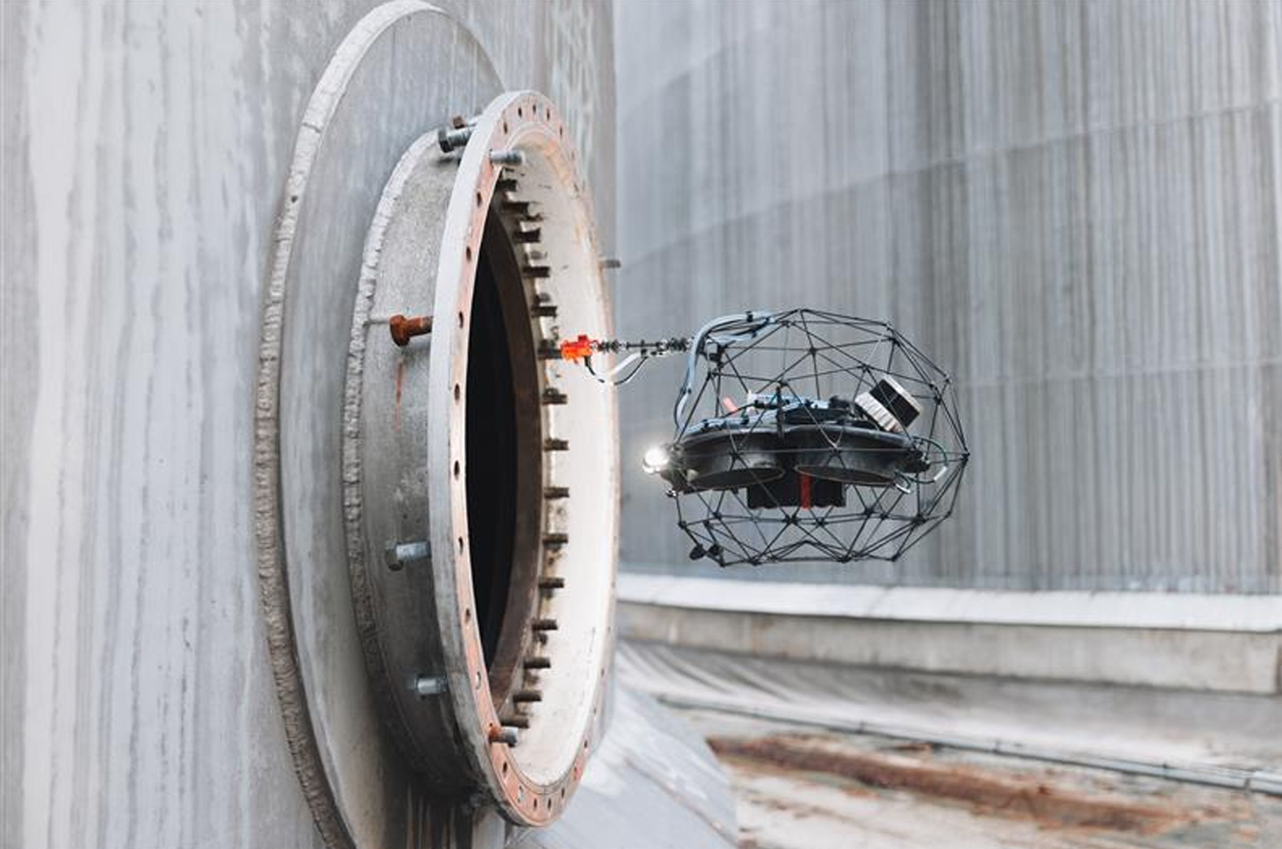 A drone with a protective cage is flying near the opening of a large industrial tank for inspection purposes.