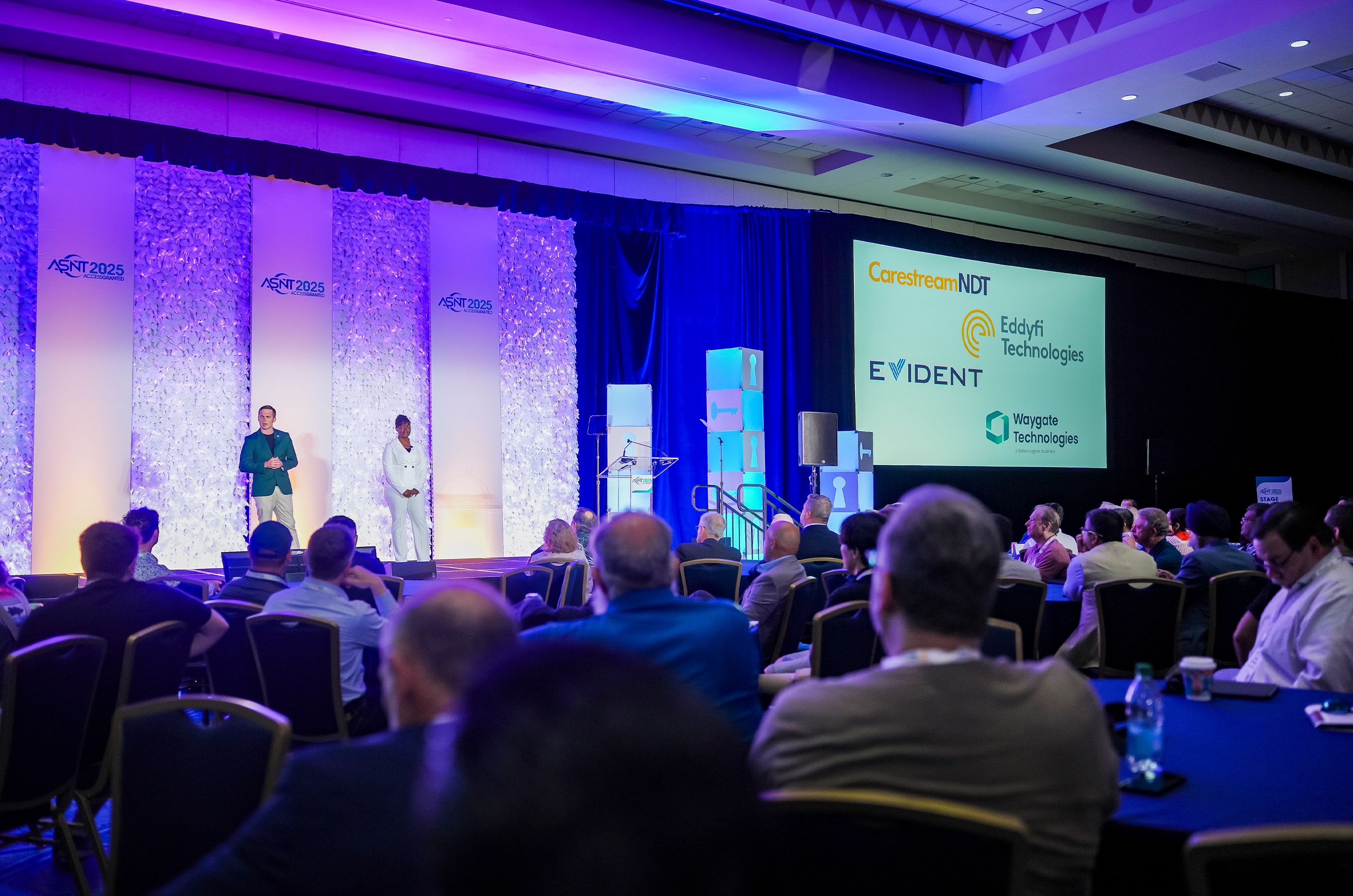 A conference room with two presenters on stage, a screen displaying sponsor logos, and an audience seated facing the stage.