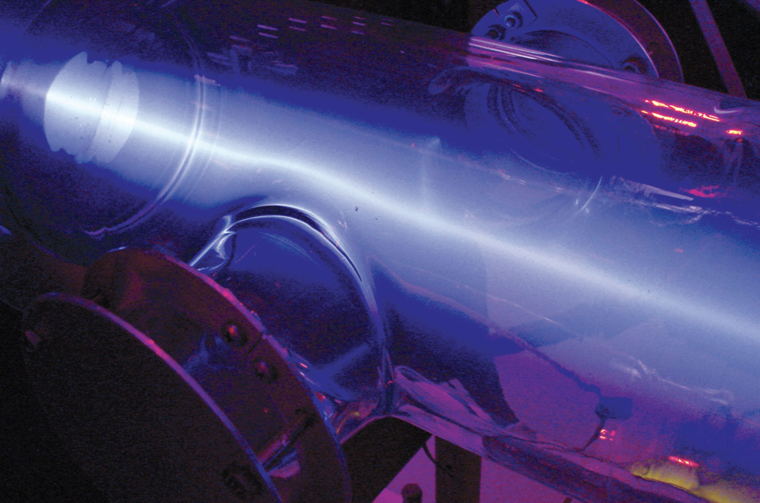 Glowing blue plasma inside a transparent tube, illuminated under purple and pink light, with metal connectors visible.
