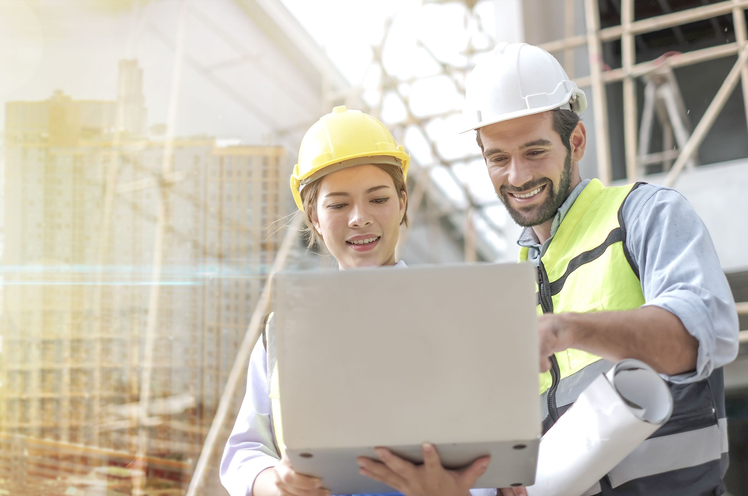 Two technicians in hard hats looking at a laptop at a building site, with scaffolding and a cityscape in the background.