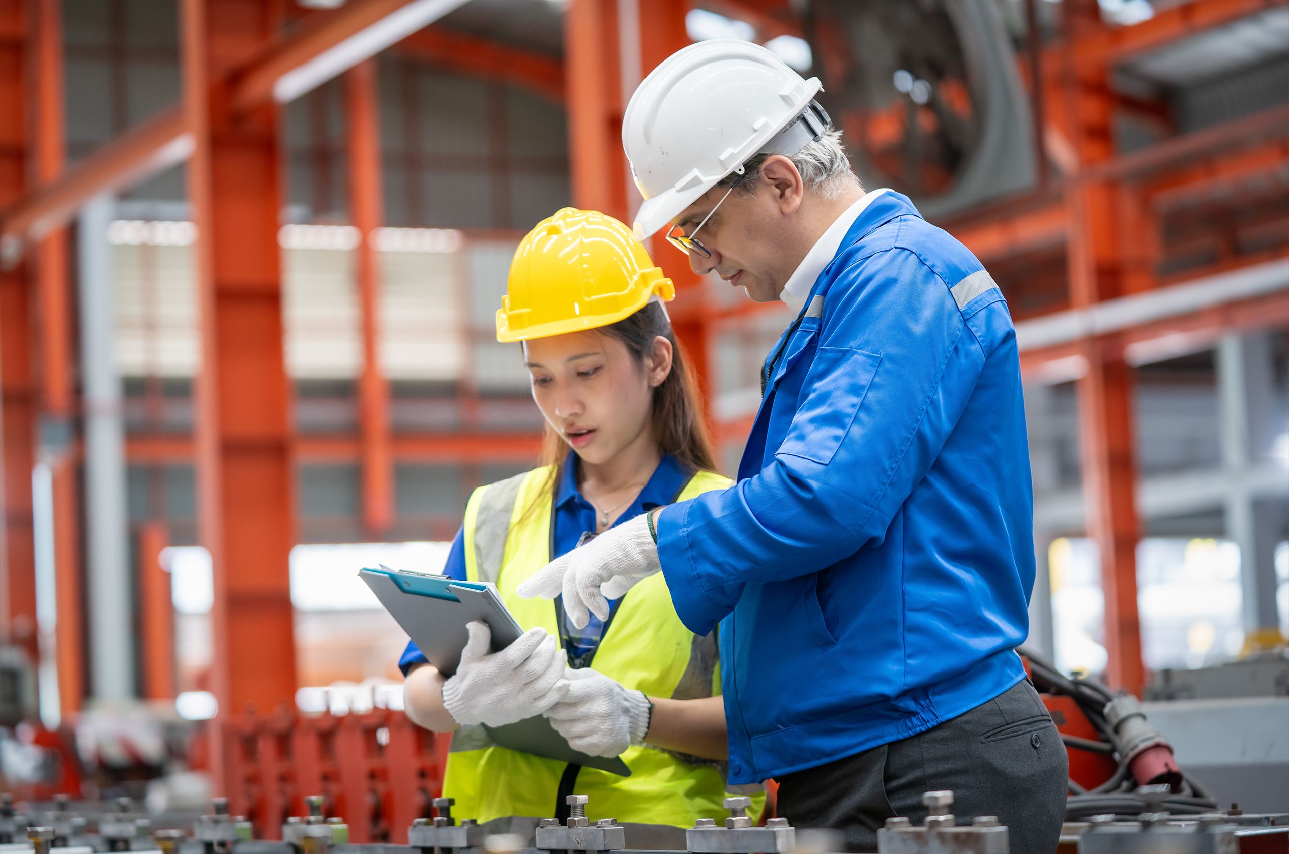 Two workers in protective gear discuss over a clipboard in a factory setting. One wears a yellow hard hat, the other a white hard hat and glasses.