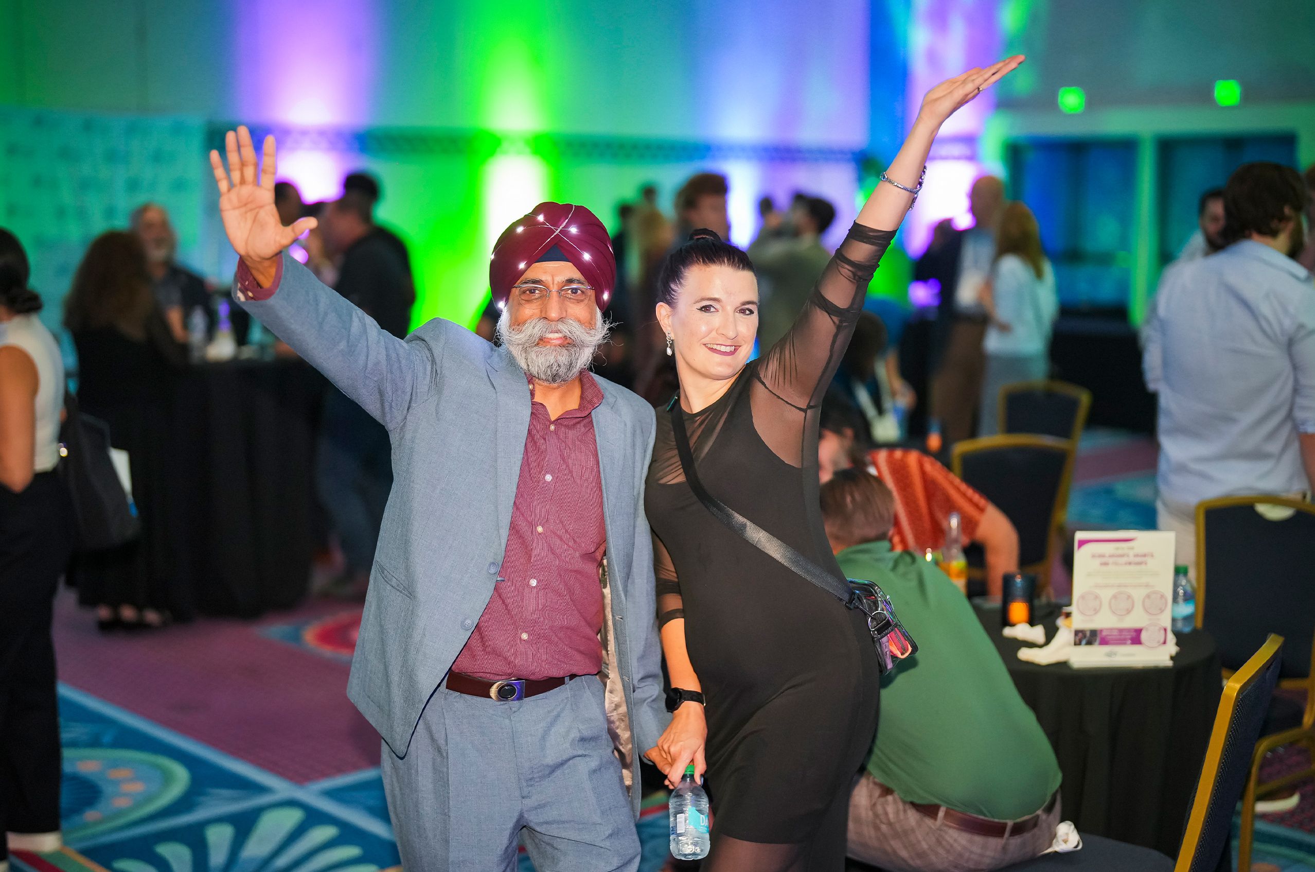 A man in a suit and a woman in a black dress pose with raised arms at a lively indoor event with colorful lighting.