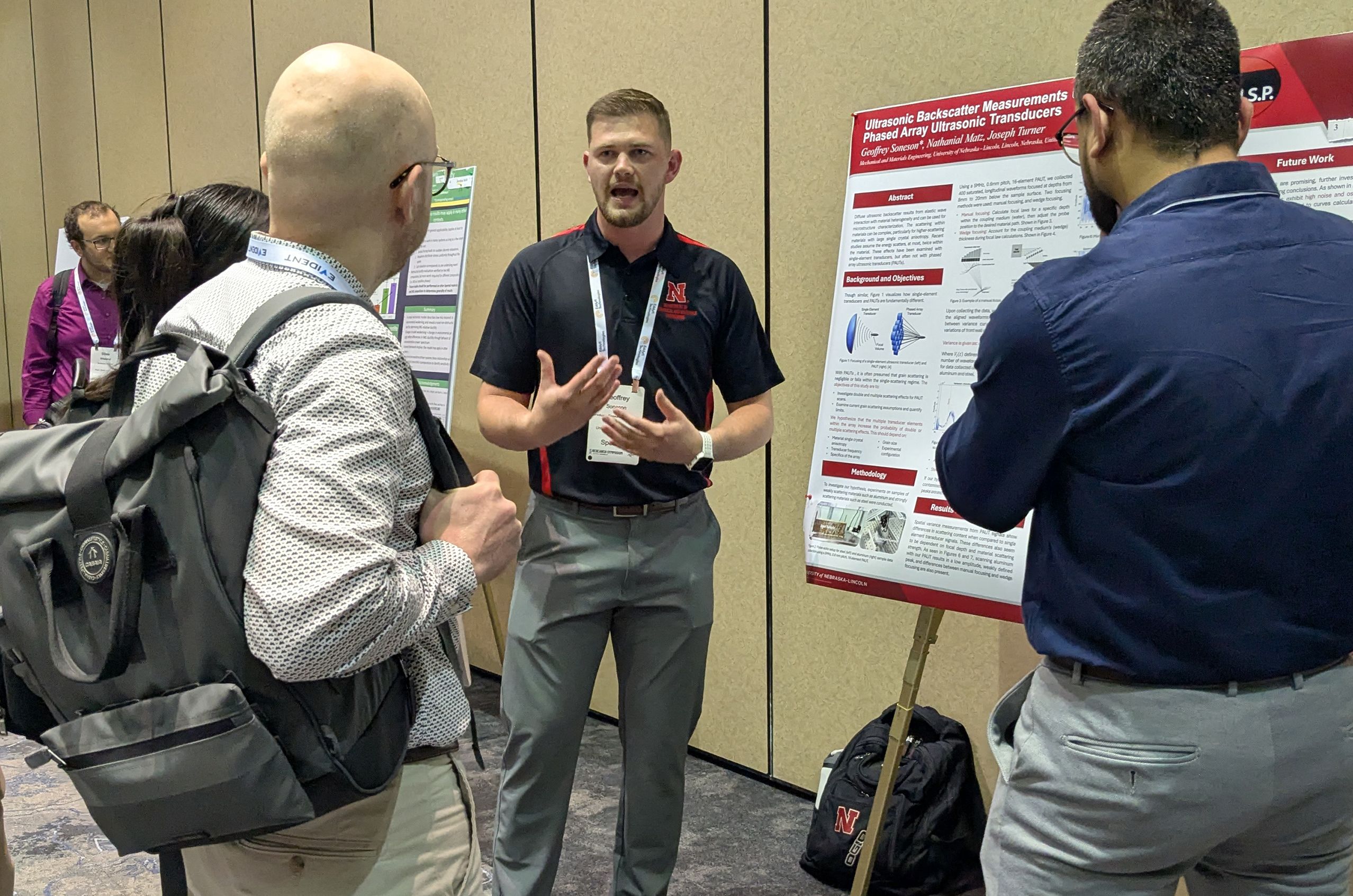 Man in black polo presenting a research poster to three attendees at a conference, gesturing while they listen.