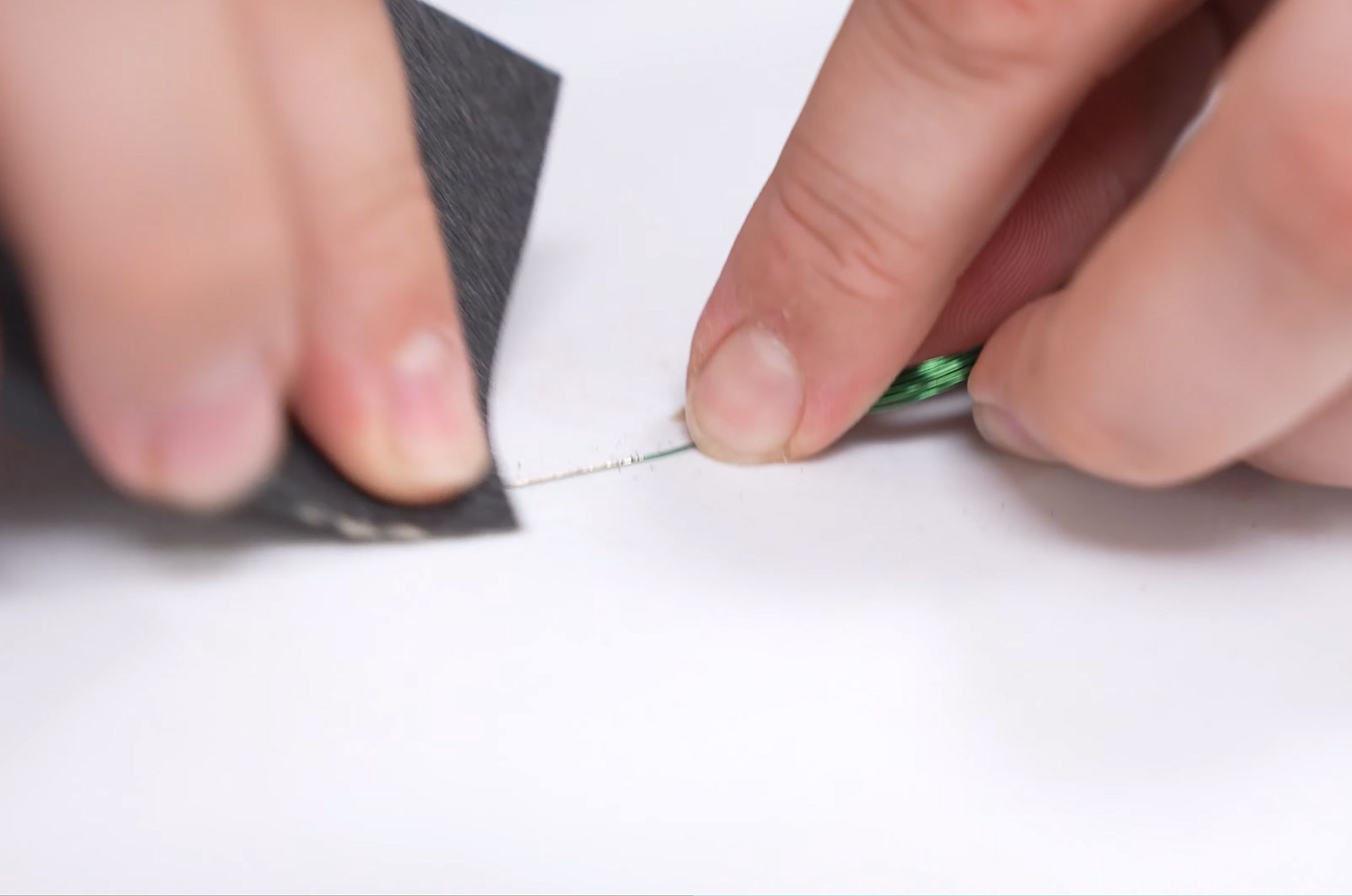 Close-up of hands sanding a thin wire with sandpaper on a white surface.