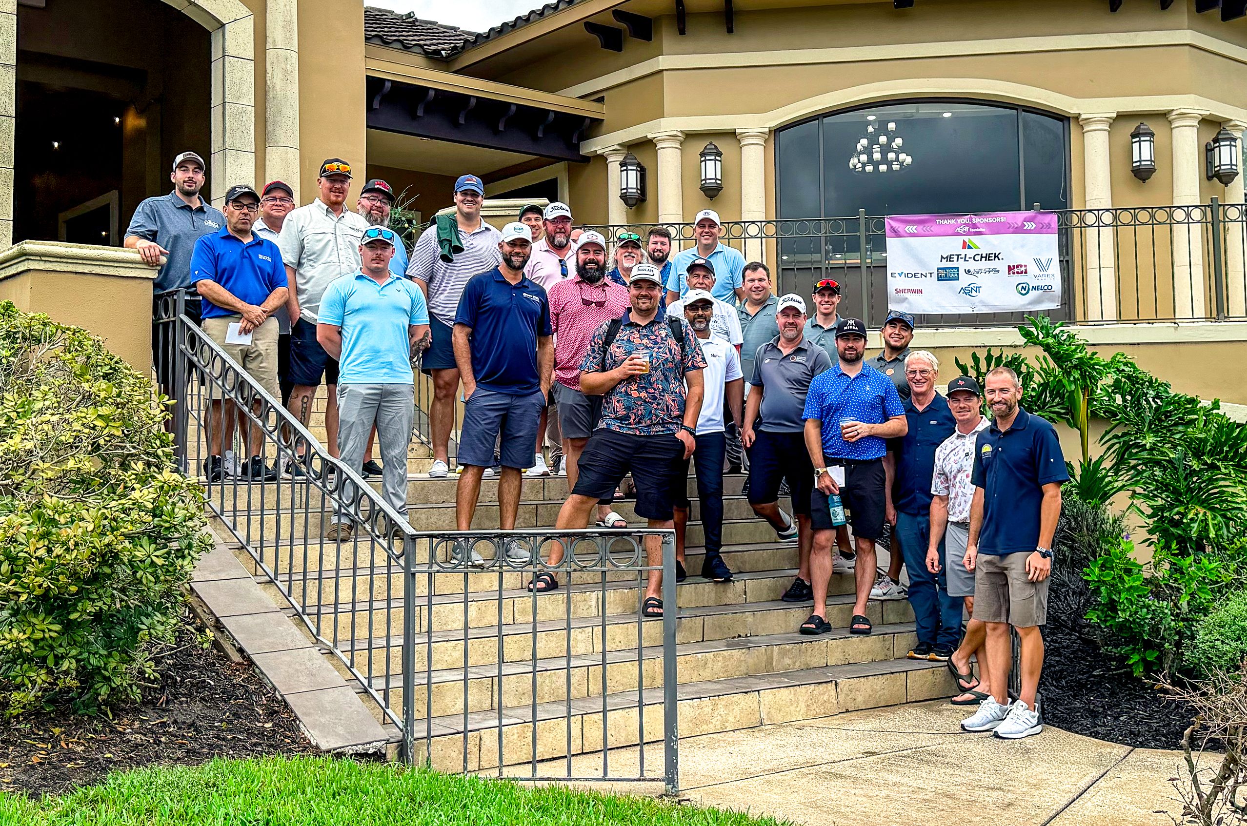 A group of men pose on steps outside a building with a banner above. Many wear casual or golf attire, smiling and standing close together.