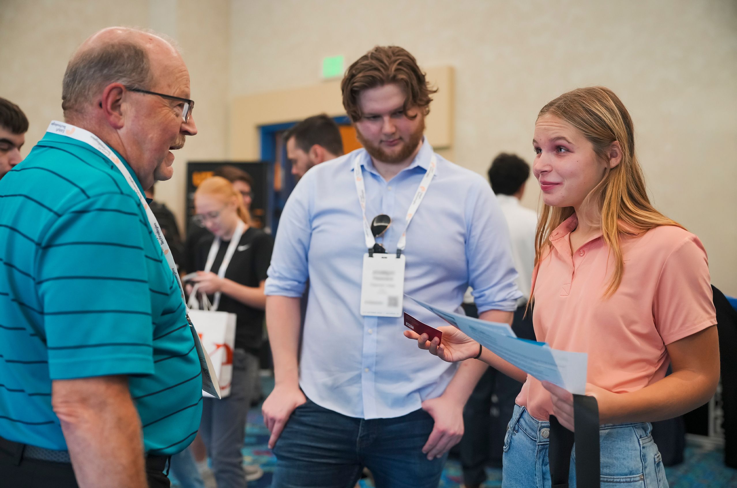 Three people in a lively discussion at a conference, wearing name tags, surrounded by other attendees in a brightly lit room.