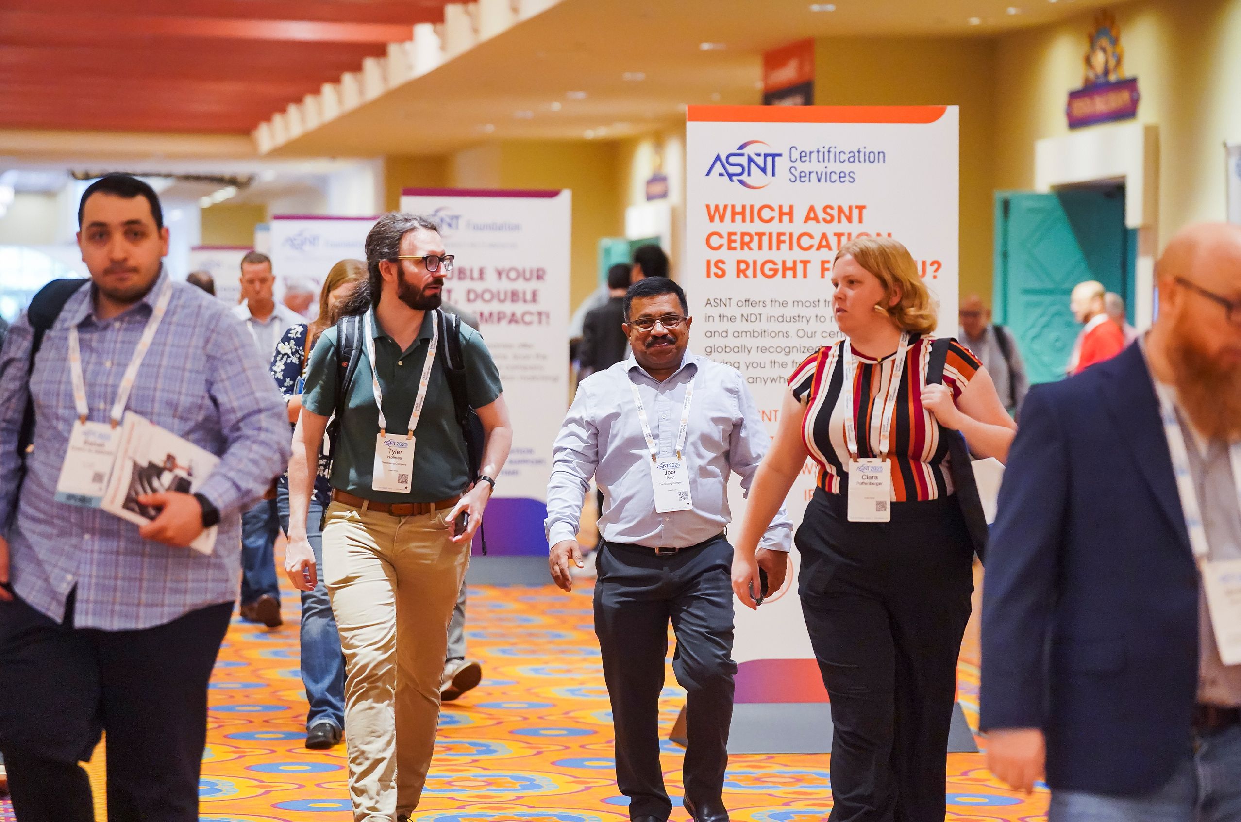 A group of people walking in a convention hall, with ASNT Certification Services signage in the background.