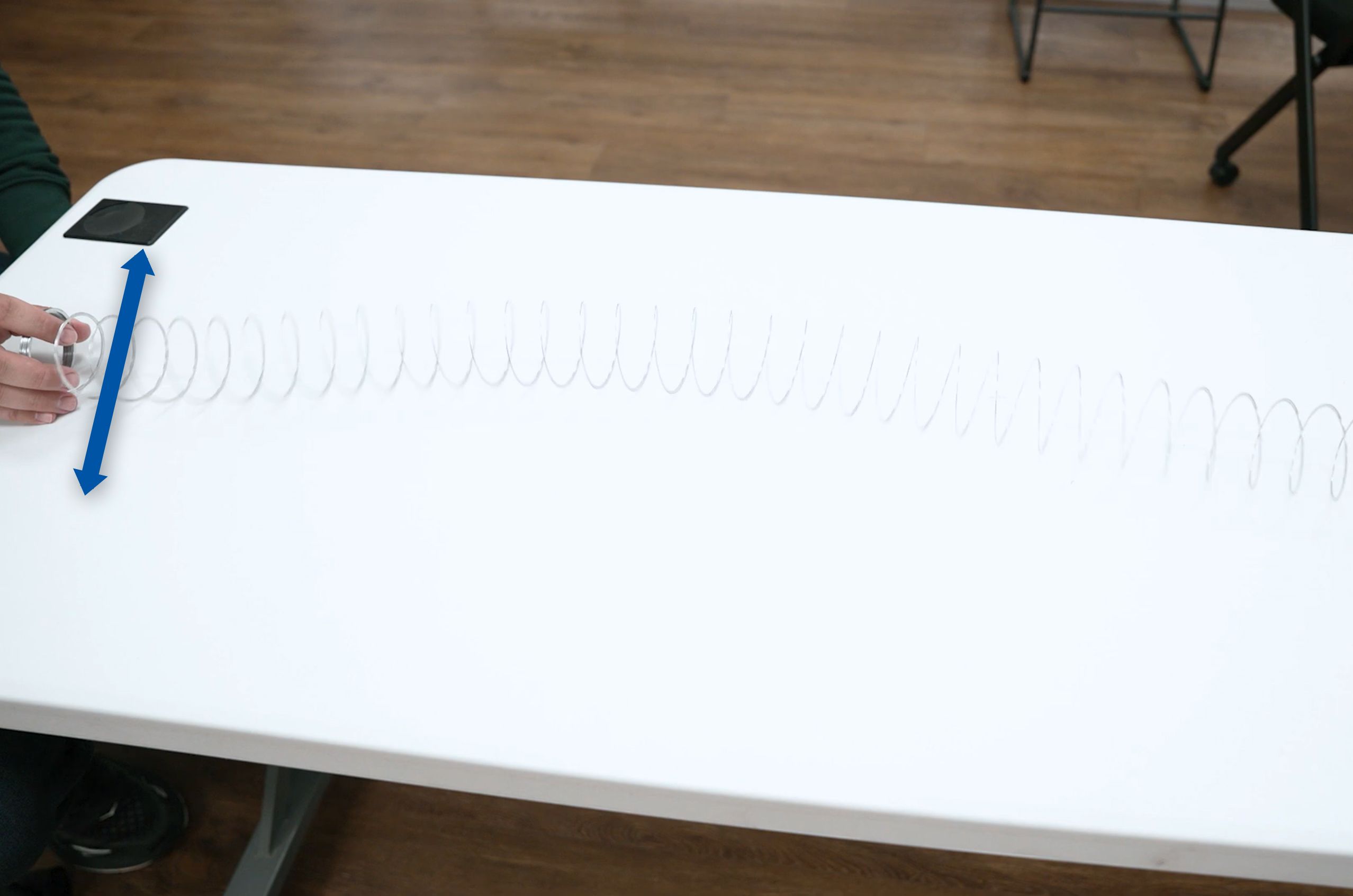 A hand holding a slinky stretched across a white table, with a blue arrow pointing showing the sideways motion put on the slinky.