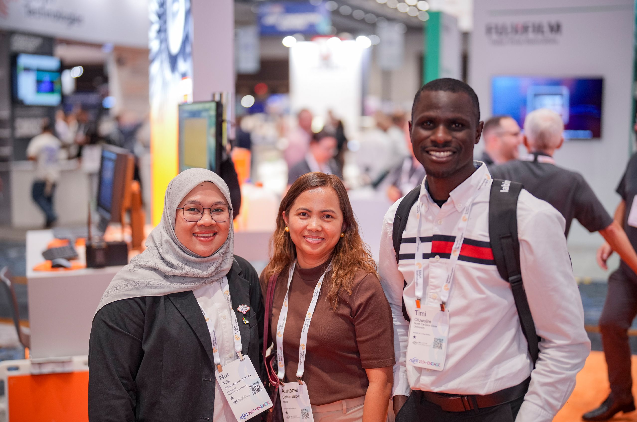 Three ASNT conference attendees smiling and posing together on the event floor, with exhibition booths and other attendees visible in the background.