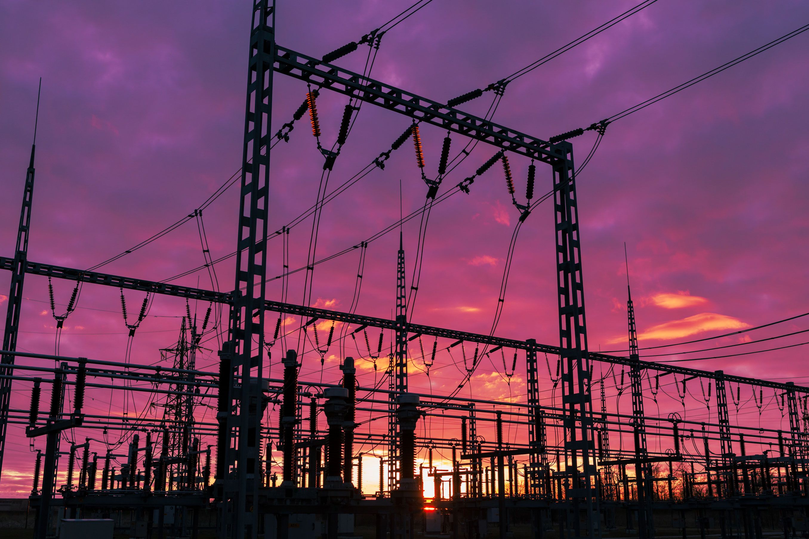 Silhouetted electrical substation towers and high-voltage lines against a vivid purple and orange sunset sky