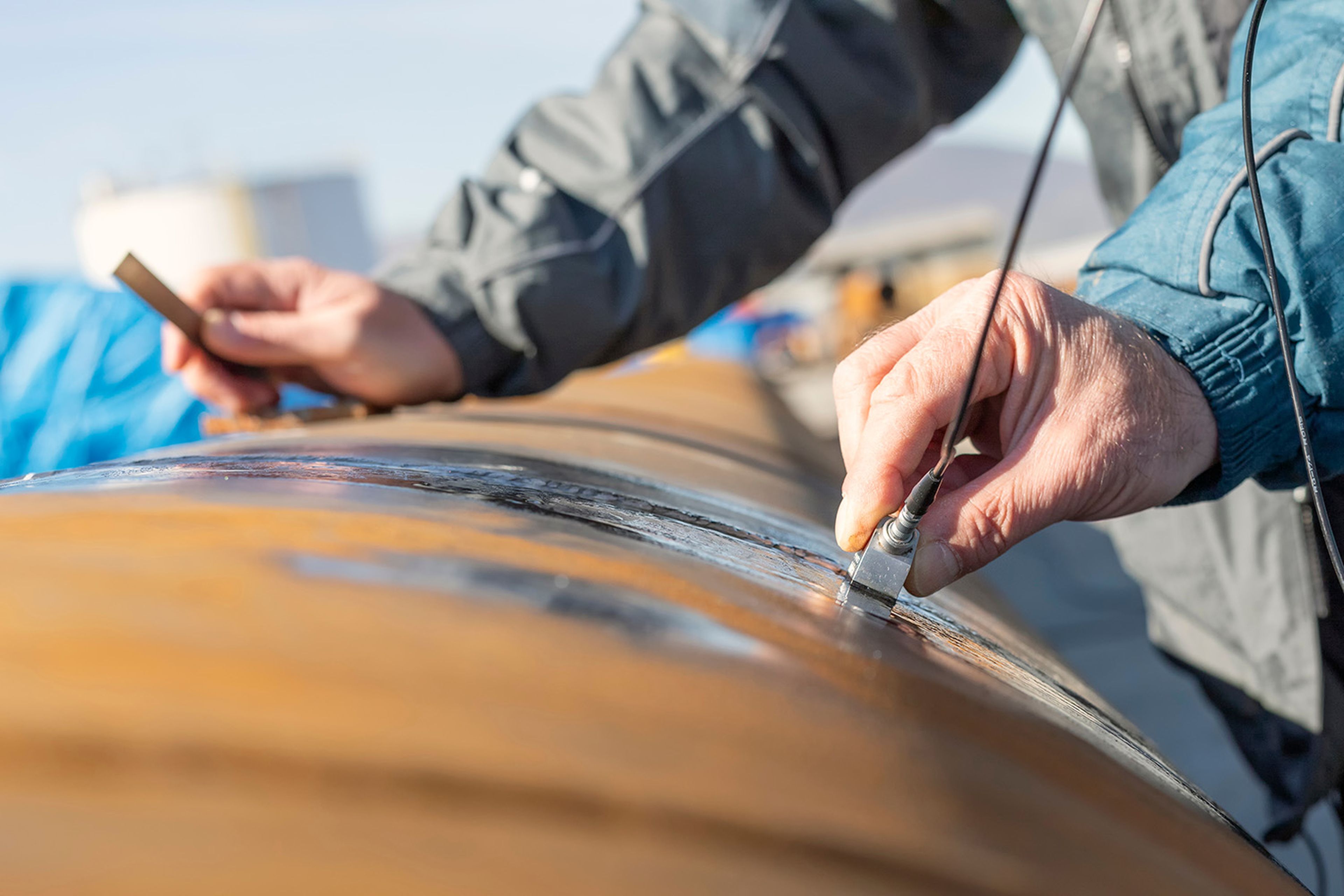 Technician using a handheld eddy current probe to inspect a weld seam on a steel pipe at an outdoor industrial site.