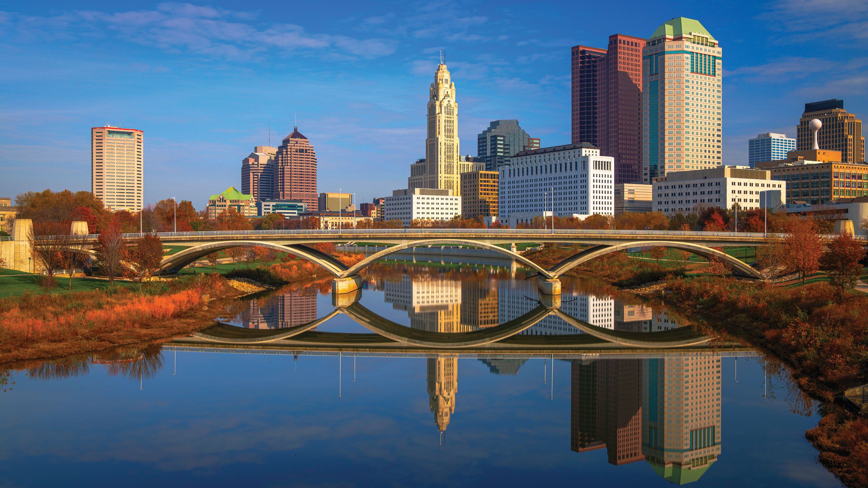 Photograph of the downtown Columbus, Ohio skyline at dusk, with office towers and the LeVeque Tower reflected in the Scioto River beneath an arched bridge framed by autumn foliage.