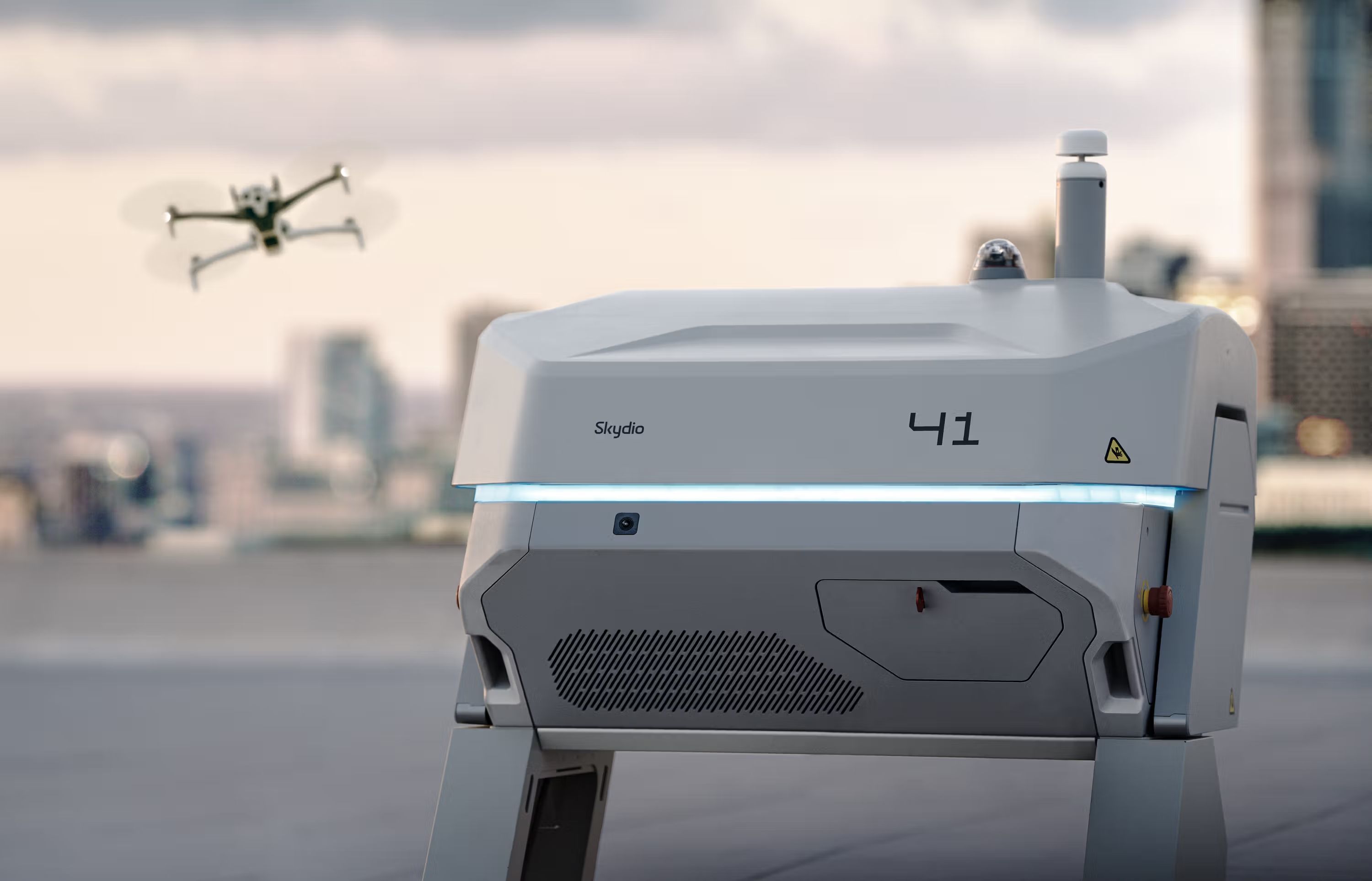 Skydio X10 autonomous drone dock station on a rooftop with a drone launching in the background against a city skyline