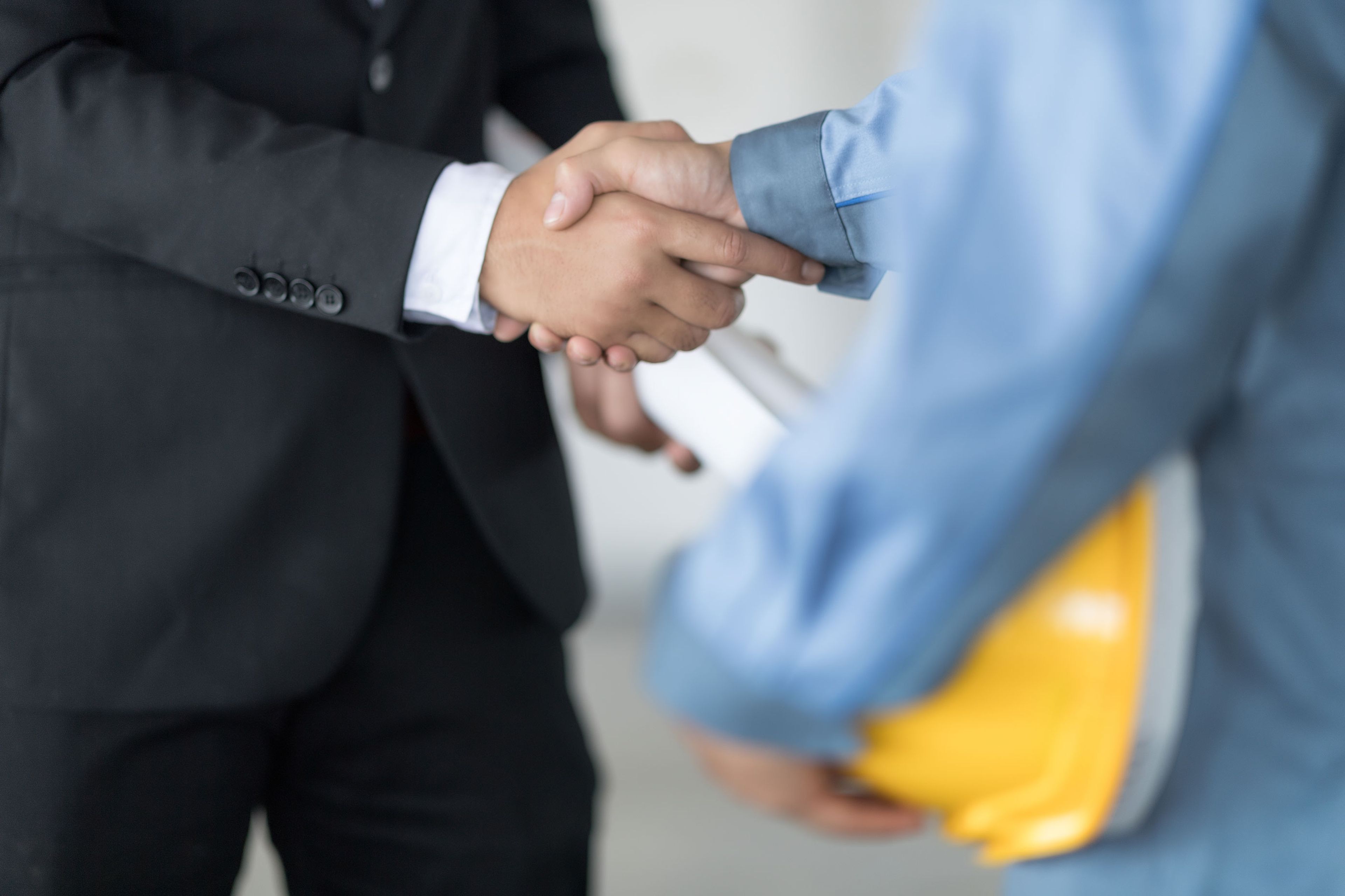A businessman in a suit shakes hands with a worker holding a yellow hard hat, symbolizing an industry partnership.
