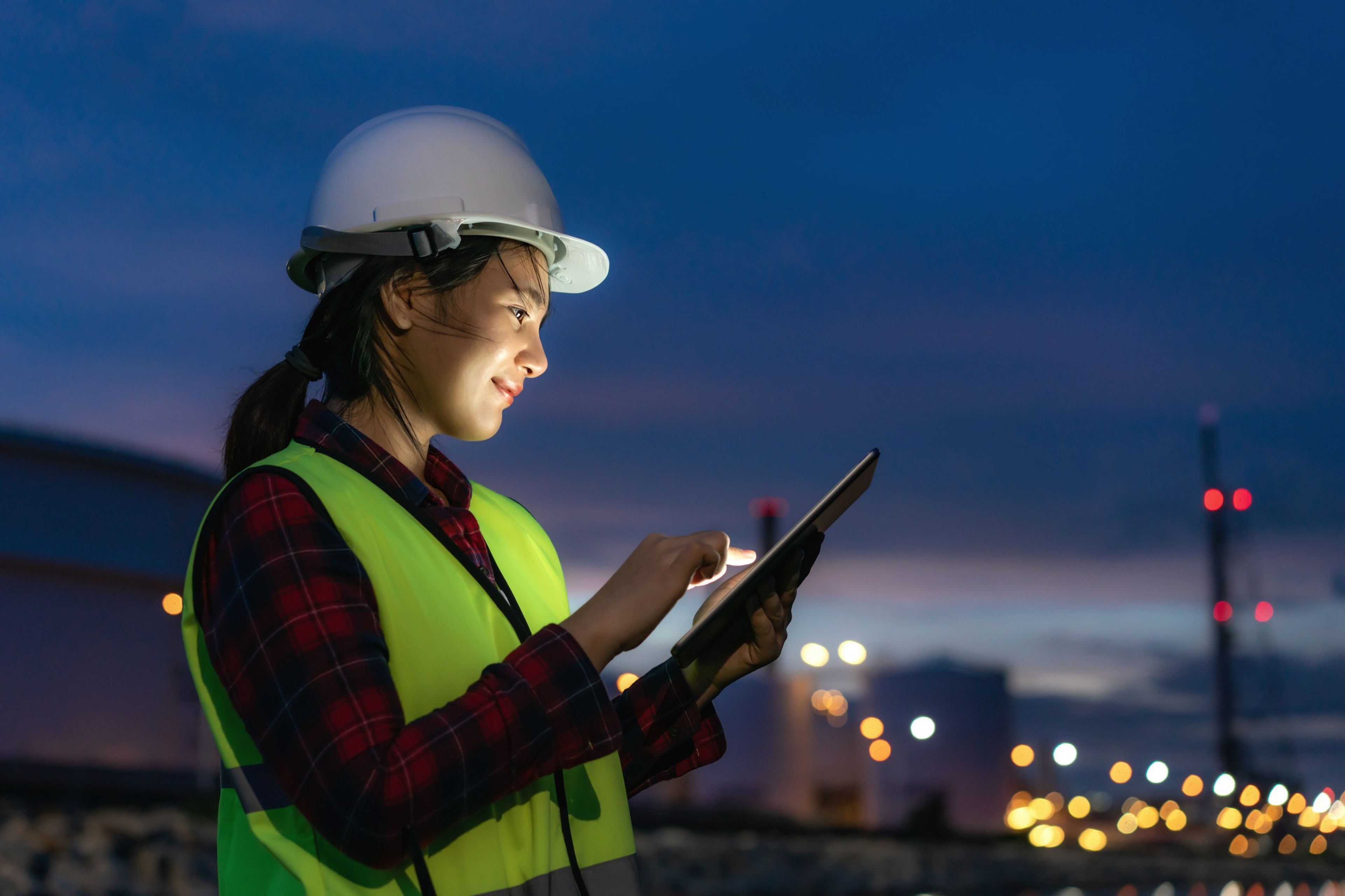 An inspector in a hard hat and high-visibility vest uses a tablet at an industrial facility at dusk.
