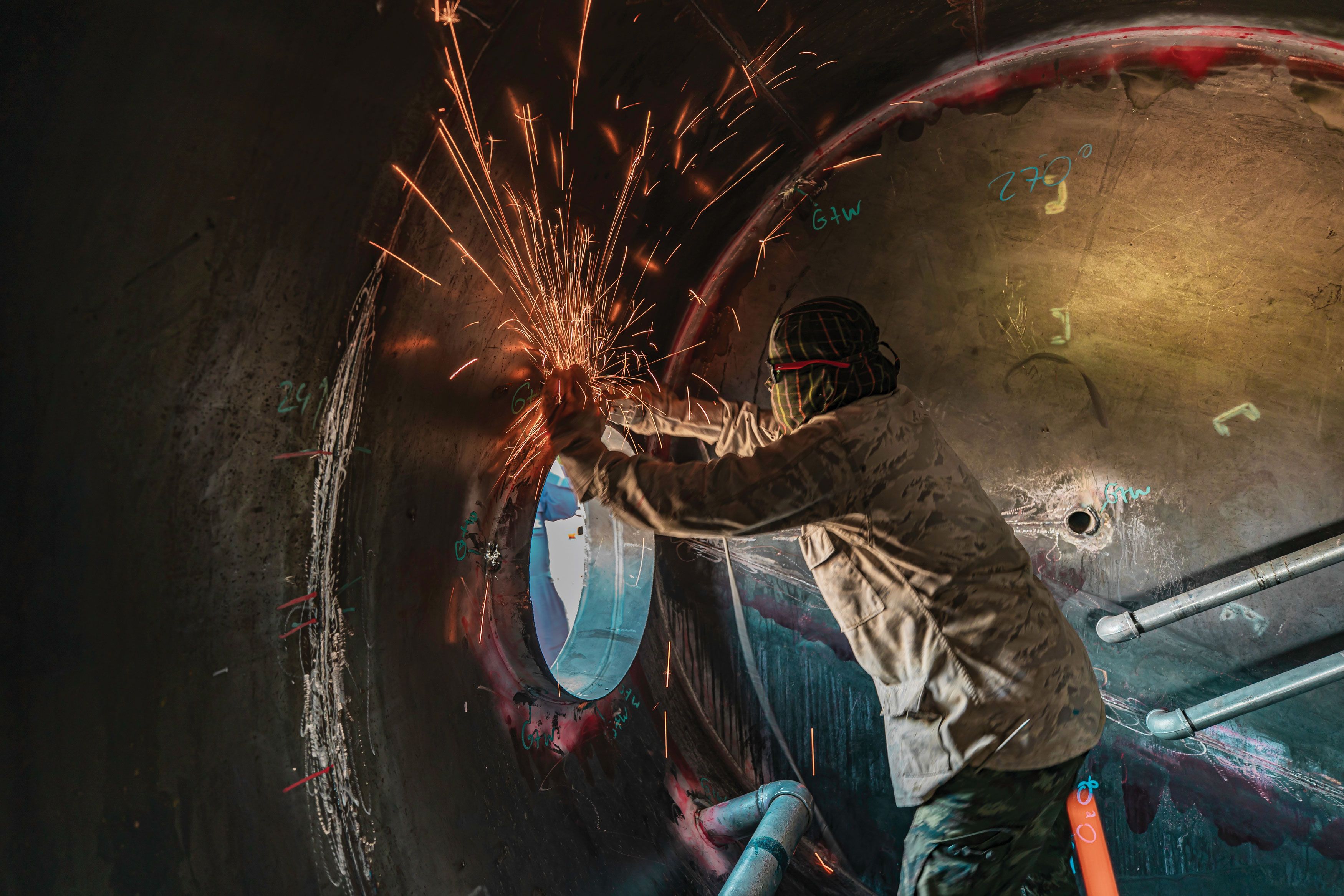 Worker welding inside a large metal cylinder, sparks flying, wearing protective clothing and headgear, with tools and markings around.