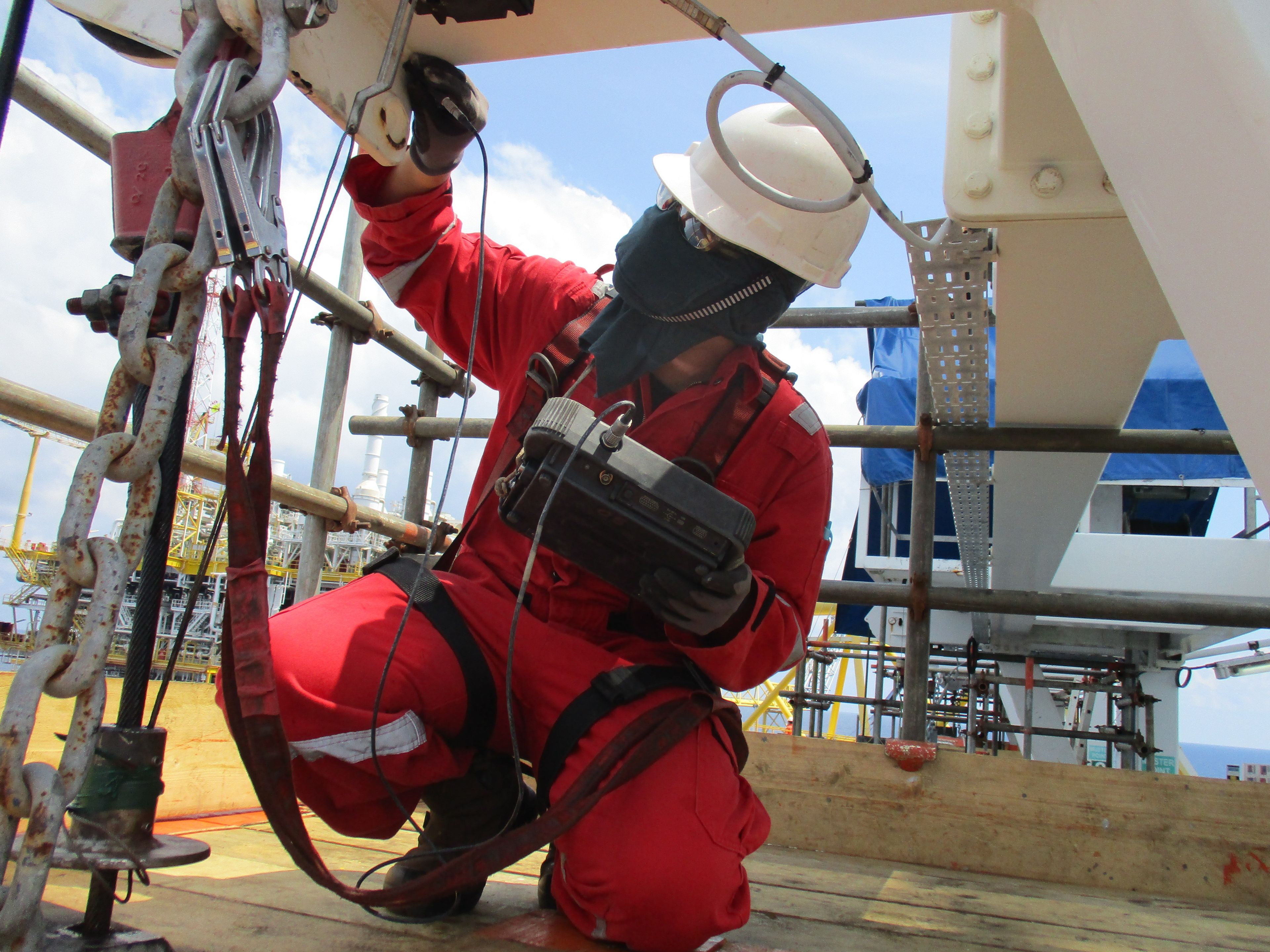 Technician in red coveralls and hard hat performing nondestructive testing on industrial structural components at an offshore site.