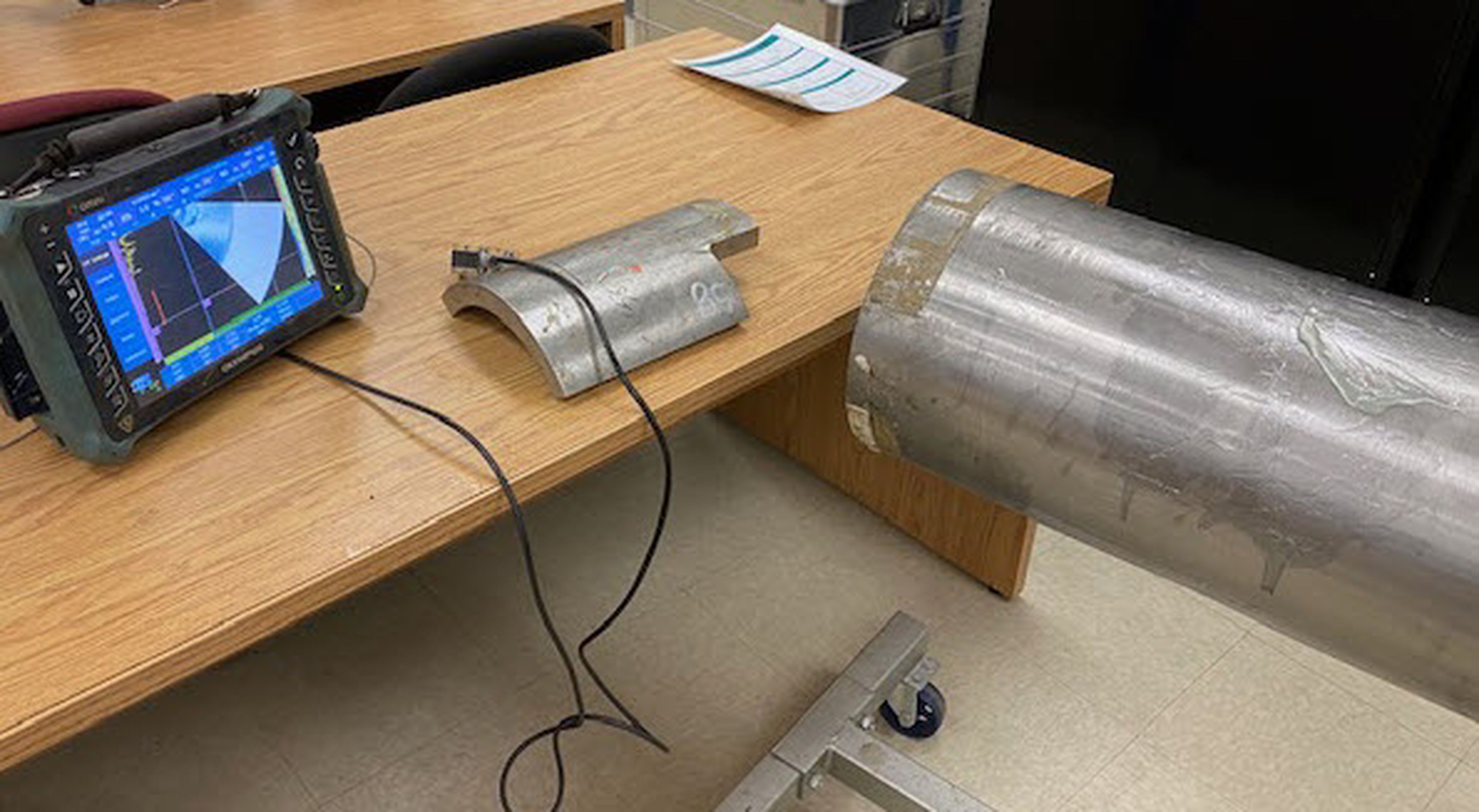 Phased array ultrasonic flaw detector displaying an S-scan, connected to a probe on a curved calibration block beside a metal pipe section.