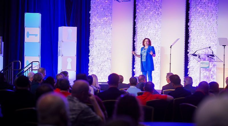 A person speaks on stage to an audience at a conference, with a blue and purple backdrop and transparent podiums.