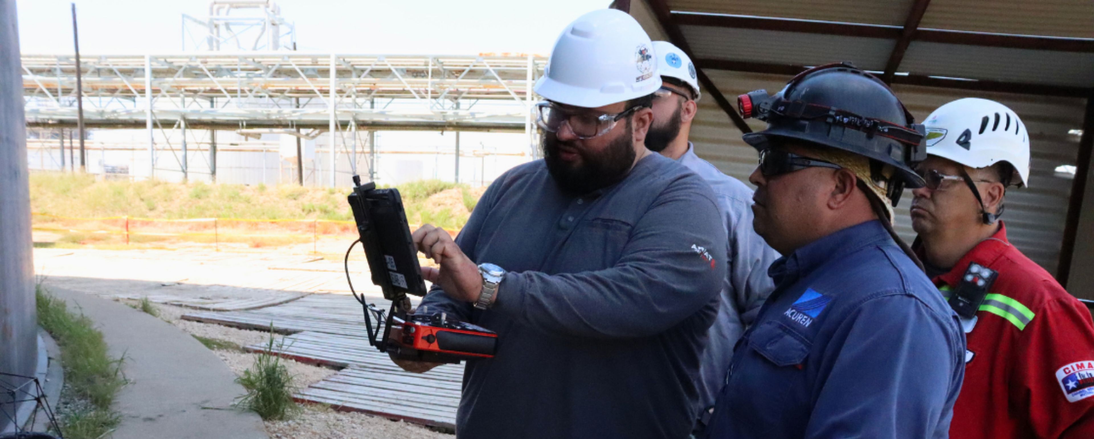 Three inspection technicians in hard hats reviewing drone flight data on a handheld controller at an industrial facility