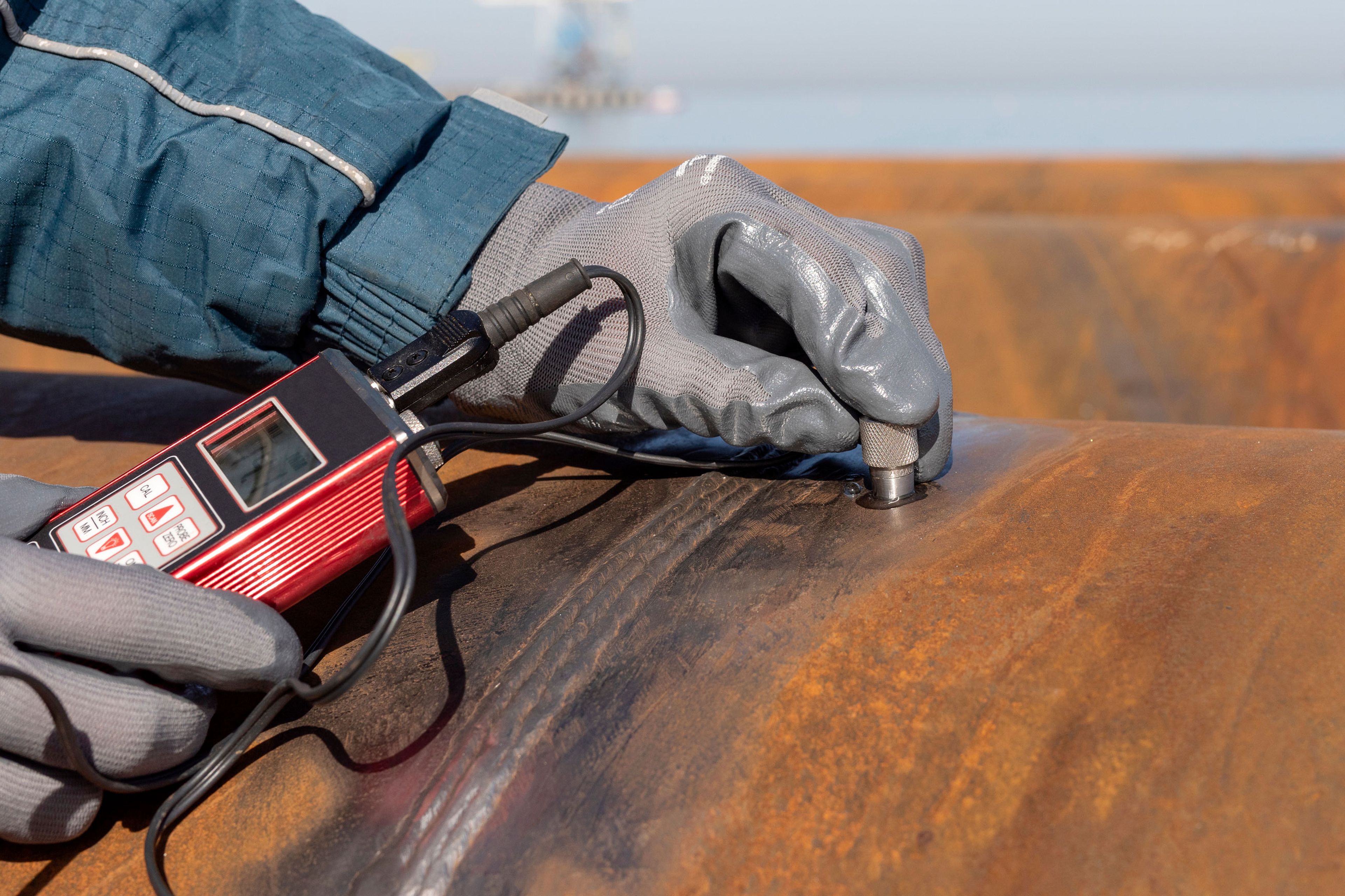 Gloved technician uses a handheld ultrasonic thickness gauge with a probe on a rusted steel surface outdoors.