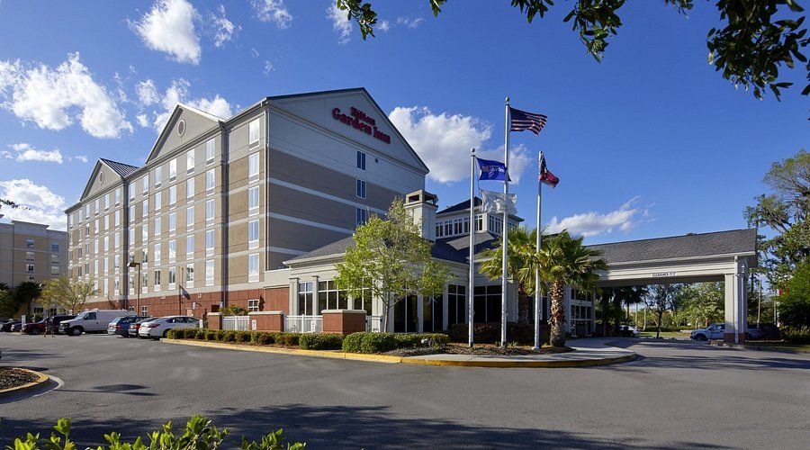 A modern hotel building with multiple floors, flags, and a tree-lined driveway under a blue sky with scattered clouds.