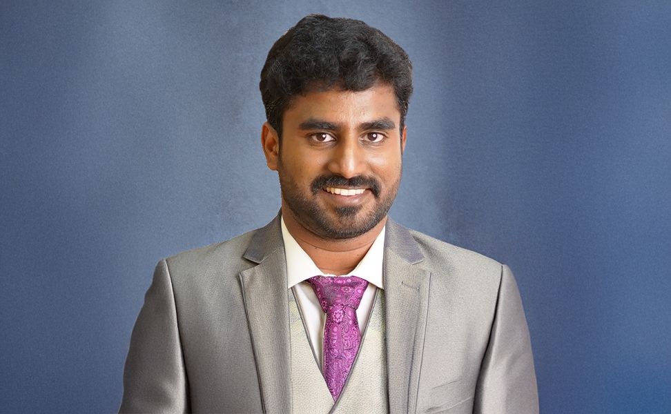 P R Dheeraj wearing a gray suit, white shirt, and purple tie, smiling against a plain blue background.