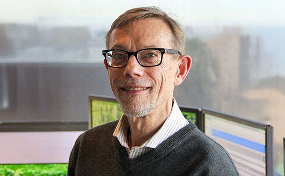 Mike Kirby smiles at the camera, wearing glasses and a sweater over a collared shirt, stands indoors, with computer monitors and a blurred window view behind him.
