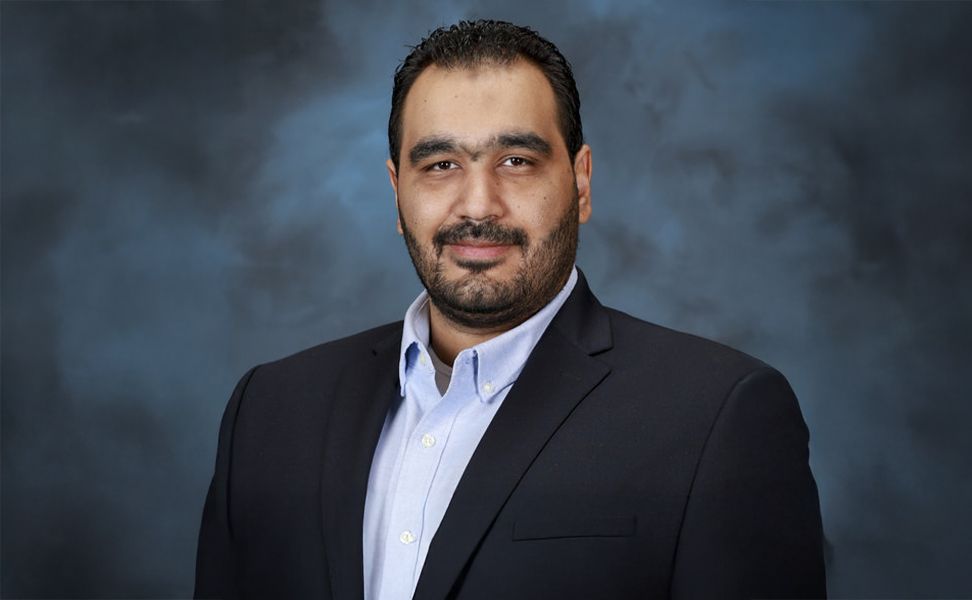 Ahmed Hassen wearing a dark suit and light blue shirt, standing against a dark, cloudy background.